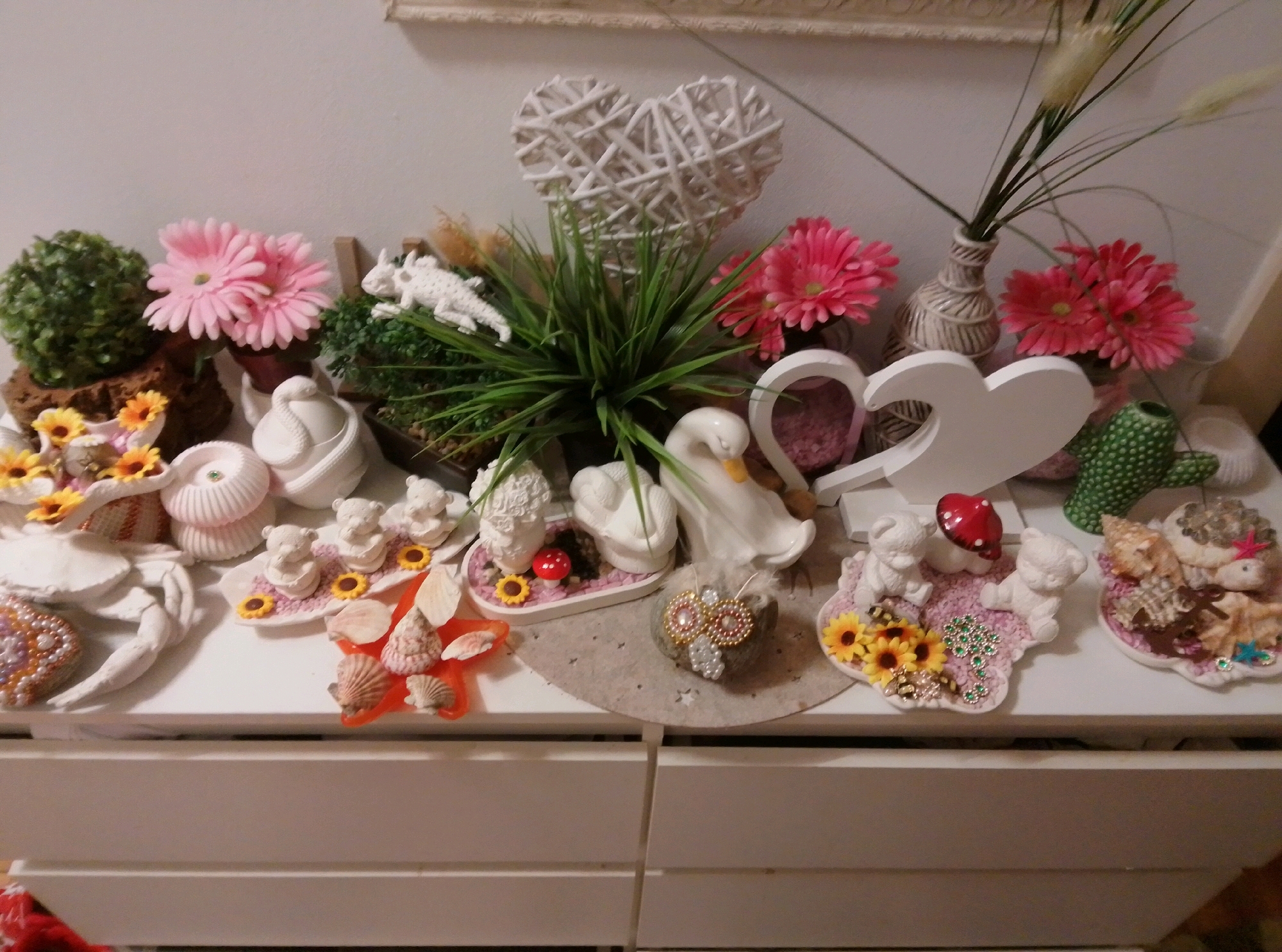 The image shows a collection of decorative items displayed on a white surface, likely a dresser or shelf. The items are varied and include artificial plants, pink flowers, white decorative hearts, animal figurines (bears, a swan, a white lizard-like creature), shells, and a decorative mushroom. Some of the items are arranged on small decorative mats. The background is a plain white wall. The overall impression is of a collection of charming, whimsical trinkets. There is no specific indication of the location within the city of Wedel, Germany, nor are there any people present. The scene appears to be indoors, in a well-lit environment.