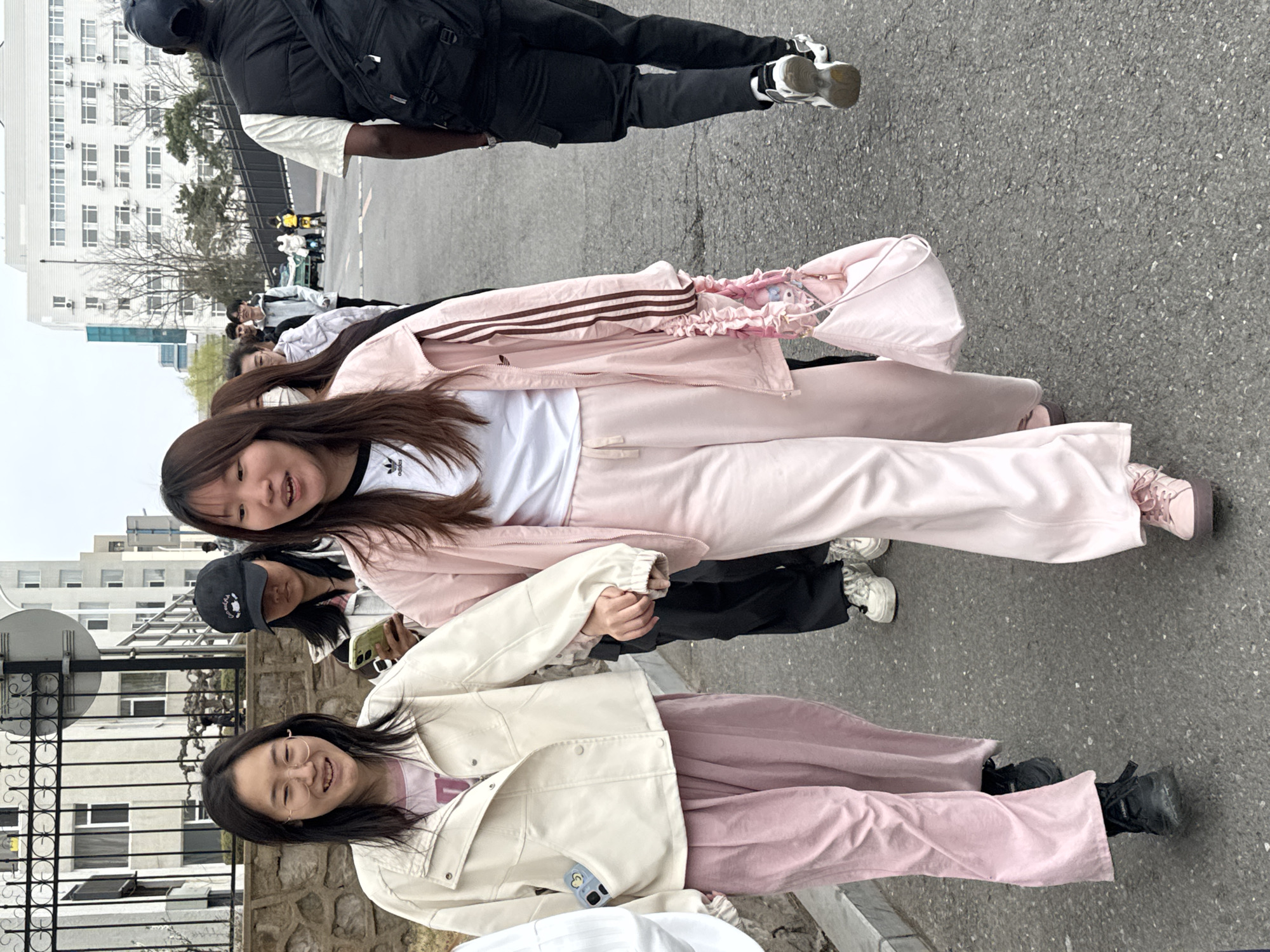 The image shows a group of young people standing together outdoors. Three girls are prominently featured in the foreground. The girl on the top is wearing a pink Adidas tracksuit. The bottom girl is wearing a pale pink jacket and mauve pants. They are holding hands. Another girl wearing a black baseball cap and black pants is partially visible between them. In the background, other people are walking. Behind them is a street in Zhongtun, China, with a building visible in the distance. The weather appears to be overcast. The ground is paved.