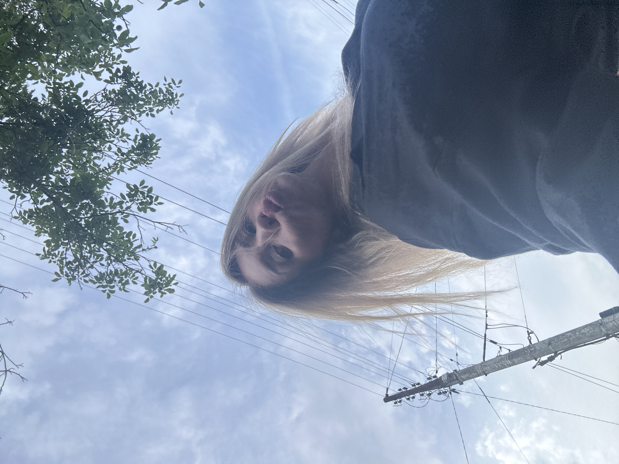 A low-angle, full shot captures a young woman from the shoulders up, outside under a cloudy sky. She has blonde hair that is being blown by the wind, and her face is angled towards the camera, with her lips pursed as if for a kiss. She is wearing a dark-colored hoodie. In the background, to the left, is a utility pole with wires extending across the frame. To the right, a portion of a leafy green tree is visible. The scene appears to be in a residential or semi-rural area, possibly in the city of Armavir, Russia.