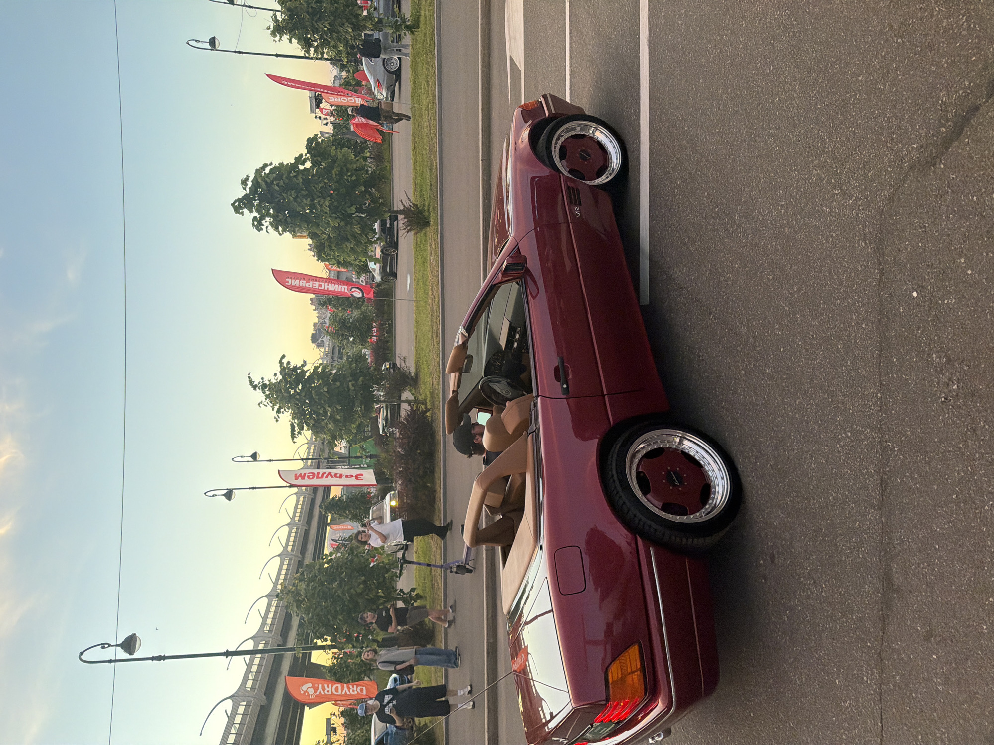 The image shows a maroon convertible Mercedes-Benz SL parked on a street. A person is seated in the driver's seat, facing away from the camera. The car has a tan interior and custom wheels. In the background, several people are standing and walking, some are holding flags. There is a pedestrian overpass in the distance, as well as trees and streetlights. The sky is a clear blue with some wispy clouds. The scene appears to be a city street during the day.