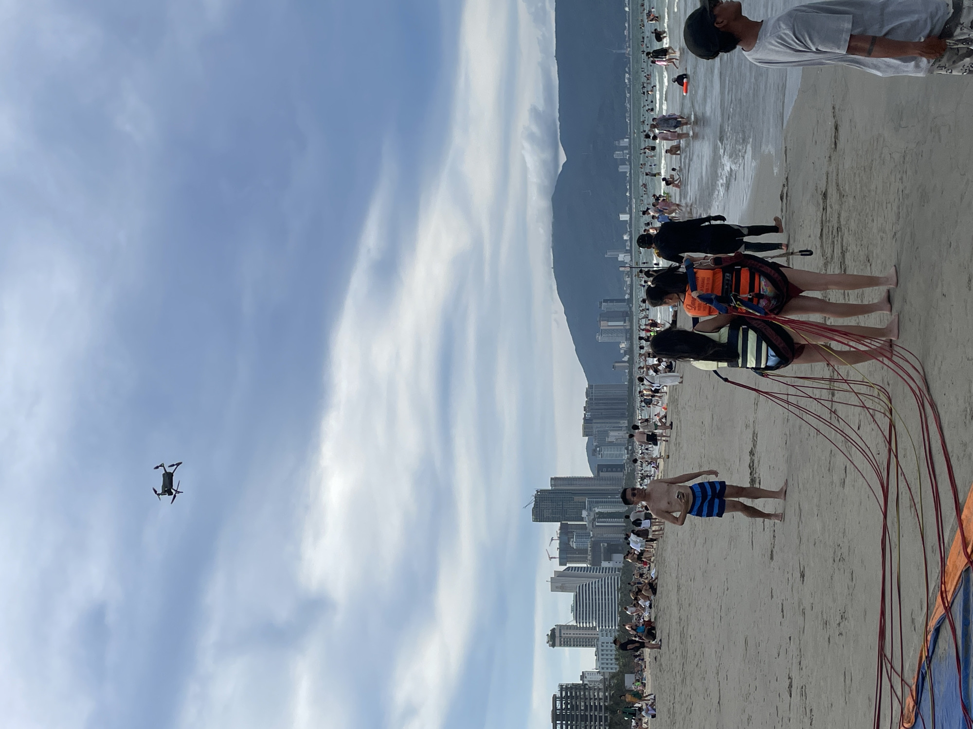 A drone flies high in a cloudy sky above a crowded beach in Đà Nẵng, Vietnam. The beach is lined with buildings and mountains in the distance. People are sunbathing, swimming, and playing in the sand and water. In the foreground, two young women wearing life vests and a man in striped shorts stand on the sand. A man in a grey t-shirt and a dark baseball cap stands facing away from the camera. The scene depicts a lively day at the beach with various recreational activities.