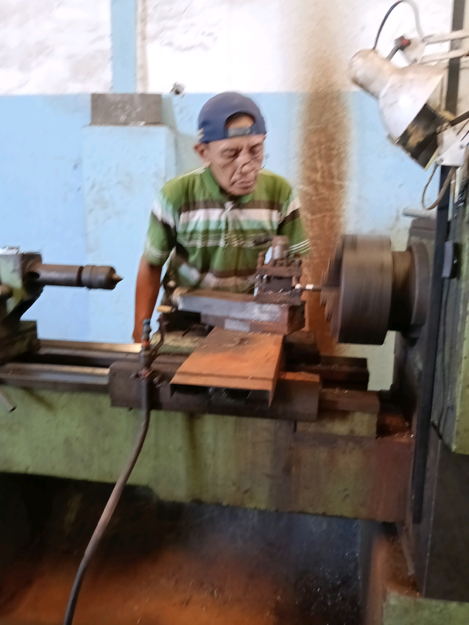 A man wearing a blue cap and a green and white striped shirt operates a lathe machine in what appears to be a workshop. He is positioned in front of the machine, with his gaze directed towards the workpiece. The lathe machine is large and appears to be made of metal, with various components such as a chuck, carriage, and tool post visible. A piece of metal is being worked on by the machine. The background shows a light blue wall, and a lamp is attached to an overhead support. The overall scene suggests a manufacturing or repair environment. The image is blurry, indicating movement or low resolution.