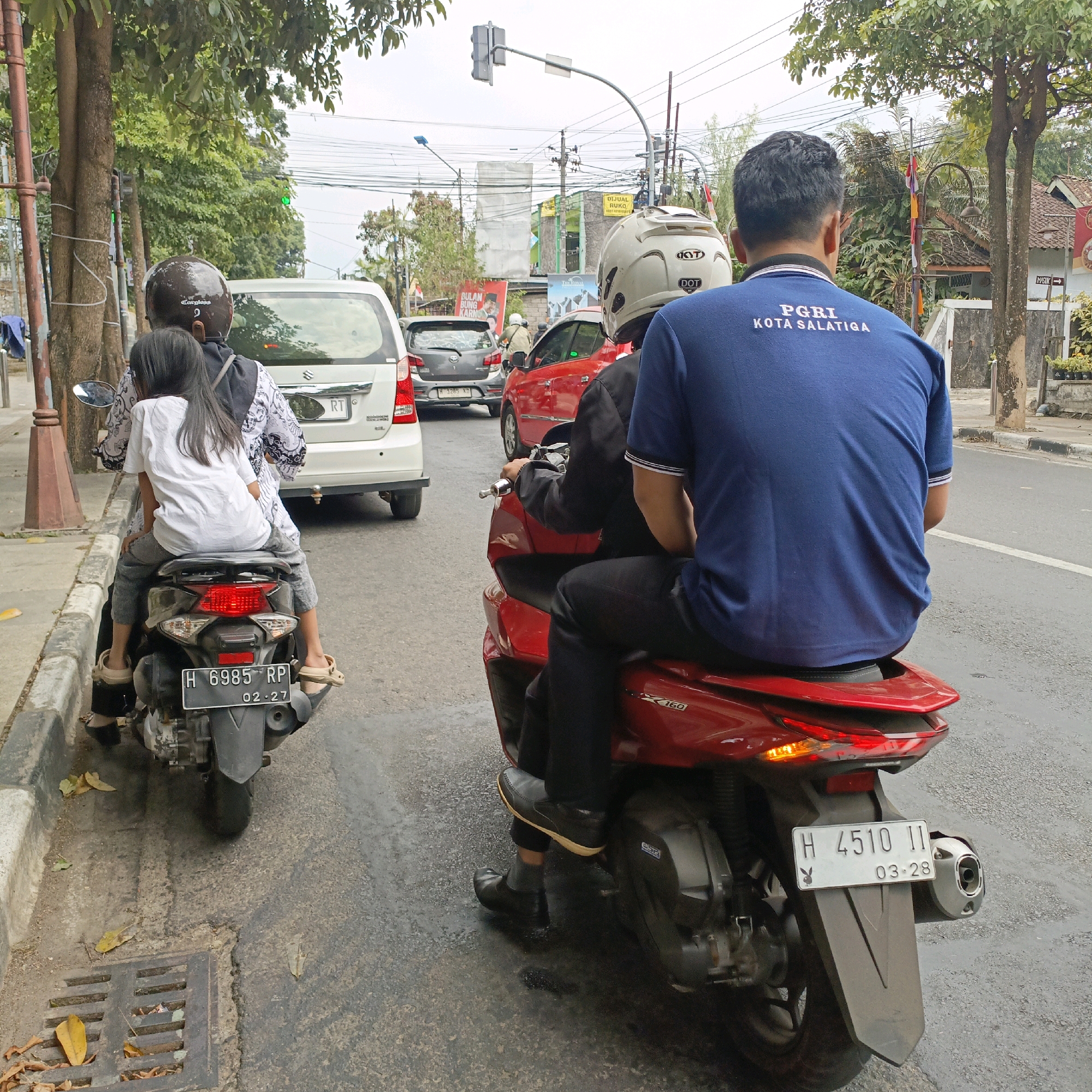 A street scene in Salatiga, Indonesia shows two motorcycles in traffic. The motorcycle in the foreground is red and has two riders. The rider in the back wears a blue shirt with the text "PGRI KOTA SALATIGA" on the back. The rider in front wears a black jacket and a white helmet. The motorcycle in the background is dark gray and has two riders, a woman wearing a patterned shirt and a child wearing a white shirt. The woman wears a dark helmet. Vehicles are stopped in traffic, including a white van and a gray car. The road is asphalt and there are trees and buildings lining the street. The sky is overcast.