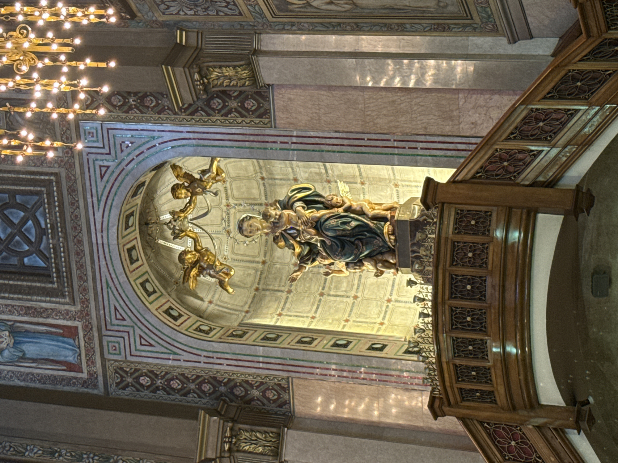 The image shows a close-up, interior shot of a religious structure, likely a church or cathedral, in Barcelona, Spain. The focal point is a statue of the Virgin Mary holding the infant Jesus, adorned in deep blue and green robes. She is depicted within a gilded, arched niche, with two golden cherubs flanking her above, one on either side. The background of the niche features a textured, illuminated wall. In the foreground, a wooden balcony or choir loft with intricate carvings and metalwork railing is visible, with decorative floral patterns. The ceiling above features a large, ornate chandelier with numerous small lights. The walls of the structure are decorated with colorful, geometric patterns and religious artwork, including what appears to be a fresco of a robed figure on the upper left. Sunlight streams in from the right, creating bright highlights on the walls.