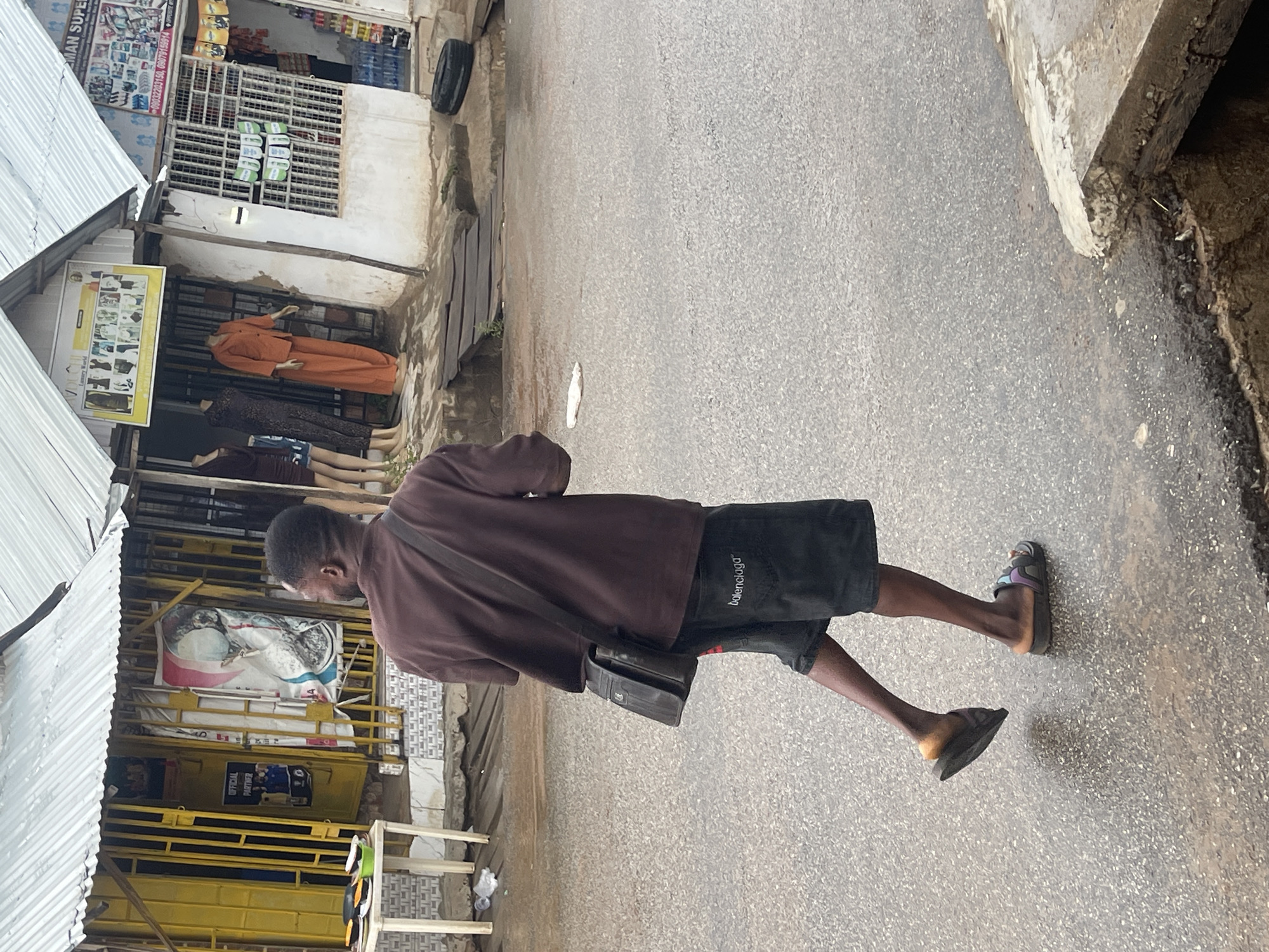 The image captures a street scene in Abuja, Nigeria, during the daytime, focusing on an individual walking away from the viewer. The person, seen from the back, is wearing a loose-fitting brown t-shirt and black shorts with "Balenciaga" written in white text on the left leg. They carry a black shoulder bag and wear dark slides on their feet; one slide has colorful details (purple, blue, green).
To the left, a series of makeshift stalls or small shops with corrugated metal roofs and yellow frames line the street. One shop features a sign that reads "AYO MAN SUPER S..." (likely Supermarket) and lists two phone numbers: "08032203150" and "08079159914". Below this, another large yellow sign prominently displays "Luxury World" alongside images of various clothing items. Inside this shop, mannequins are visible, dressed in different outfits, including a bright orange two-piece set and a patterned dark dress. Products are stacked on shelves within the shops.
The person is walking on a textured, grey asphalt road. In the upper right corner, a black tire is leaning against a wall. The scene depicts a bustling commercial area, characteristic of a local market or street vendor environment in a city like Abuja. The bright lighting suggests it is day, and there are no discernible emotions from the individual, who is captured in mid-stride.