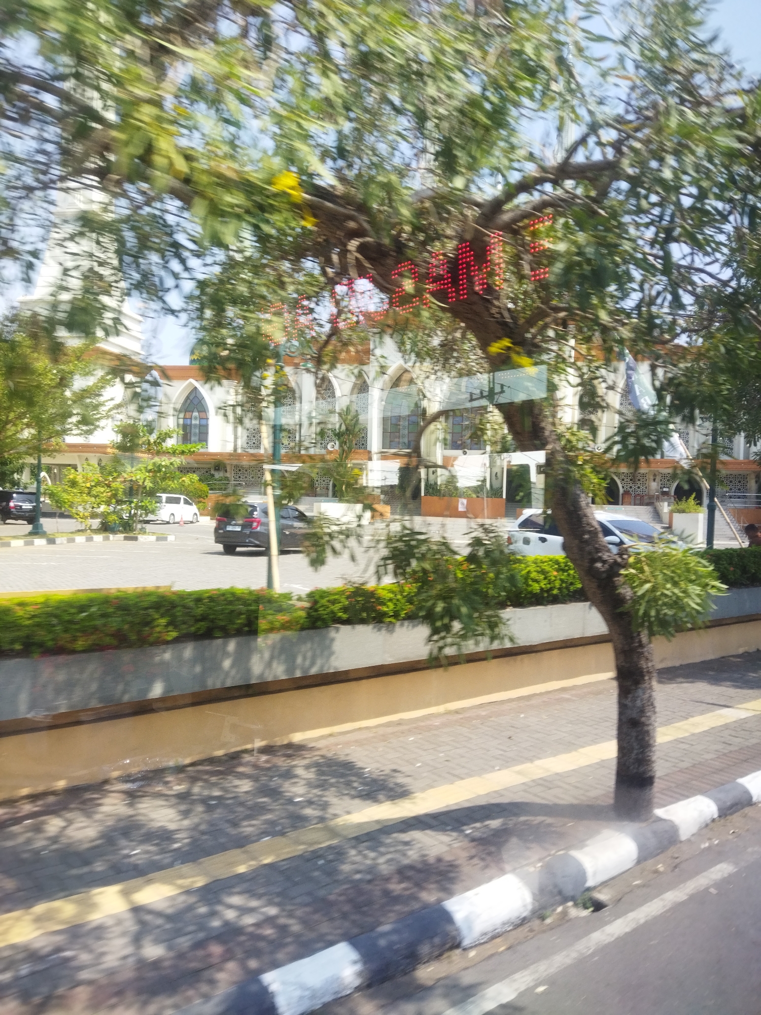 In the city of Sukoharjo, Indonesia, a blurred image captures a street scene dominated by lush green trees in the foreground. Red LED lights form a partial word, possibly "WELCOME," illuminated on the trunk of a prominent tree. Behind the foliage, a large, white building with arched windows and decorative patterns is visible, suggesting it might be a place of worship or a public institution. Several cars, including a black SUV and a white van, are parked or driving on the street in front of the building. The sidewalk features a distinctive black and white striped curb, and the street is paved with brickwork. The overall impression is a sunny day with gentle movement causing some blur in the image.