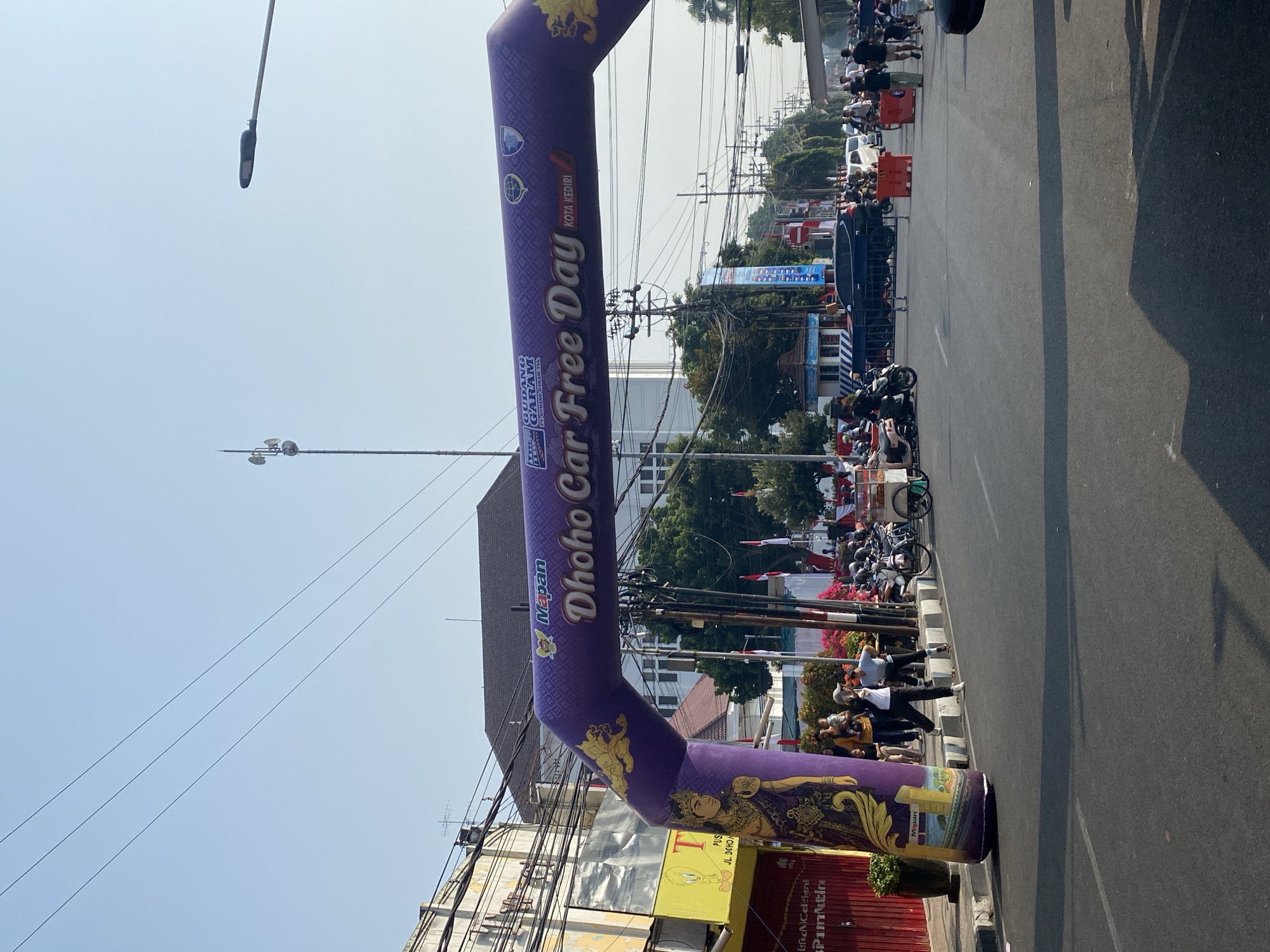 A wide-angle shot captures a vibrant street scene in Kediri City, Indonesia, on a clear, sunny day. Dominating the foreground is a large, inflatable purple archway emblazoned with "Dhoho Car Free Day" and "KOTA KEDIRI" in white lettering. The arch is decorated with intricate golden patterns and illustrations of a traditional Javanese figure on its left pillar.
The street is filled with a mix of people and vehicles, indicating an active event. Several motorcycles are parked and in motion, some ridden by individuals and others carrying passengers. A few cars are visible further down the street, suggesting a controlled traffic flow or a designated car-free zone. Pedestrians are scattered throughout the scene, some walking casually, others gathered in groups. A food cart is also present, hinting at local vendors participating in the event.
The backdrop consists of a mix of buildings, including a multi-story structure with a dark roof and a building with many windows. Electrical wires crisscross overhead, and a tall communication tower stands in the distance. The street itself is paved and lined with sidewalks. Red and white flags are displayed along the street, adding to the festive atmosphere. The overall impression is one of a lively community event taking place in an urban setting.