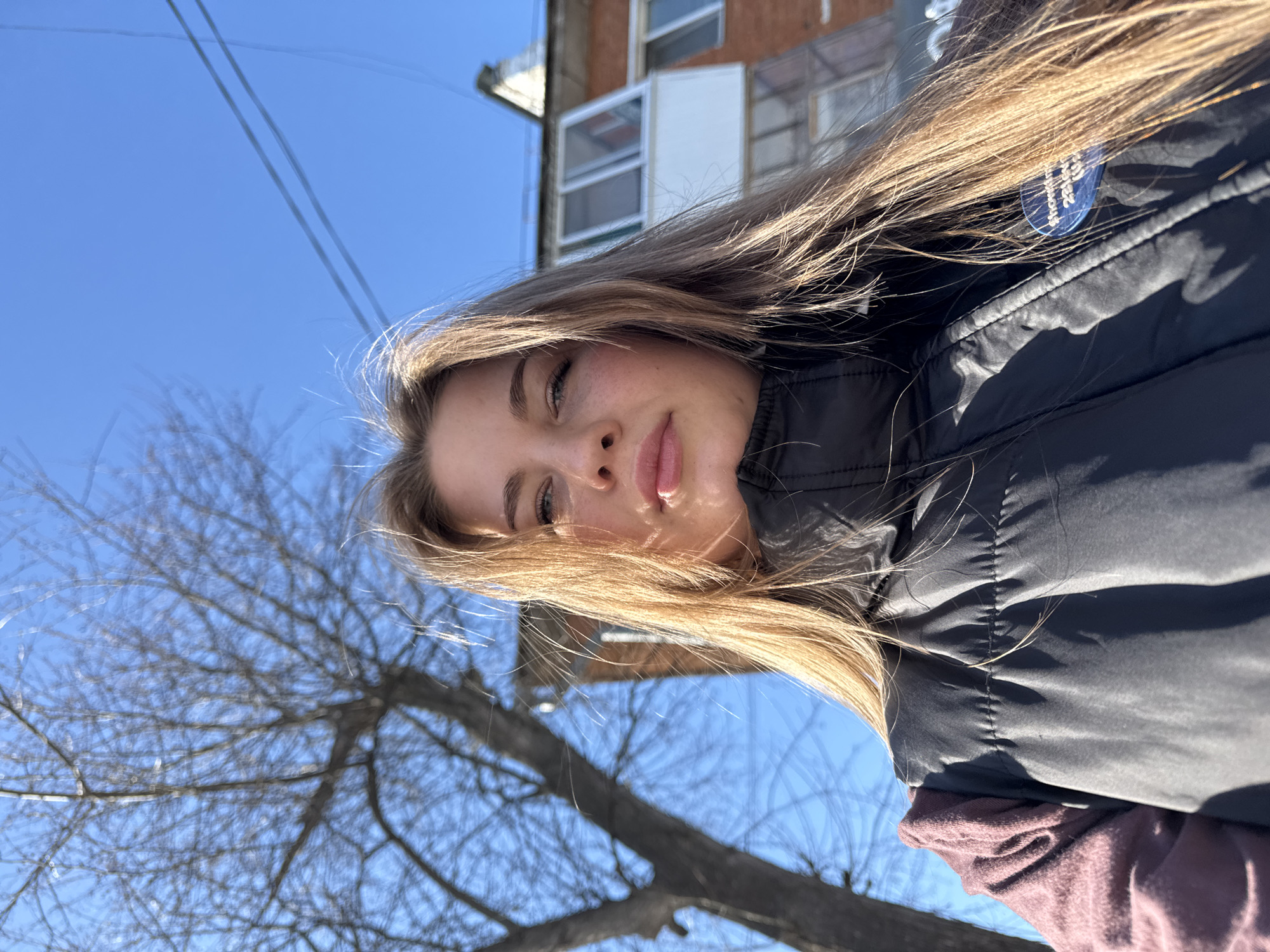 A young woman is facing the camera, tilted slightly to her right, with a blue sky and a building in the background. The woman has long, light brown hair. She is wearing a black puffer jacket over a dark pink shirt. The location is Chaikovsky, Russia, as indicated by a small round tag attached to the jacket. A bare tree is visible in the lower left corner of the frame.