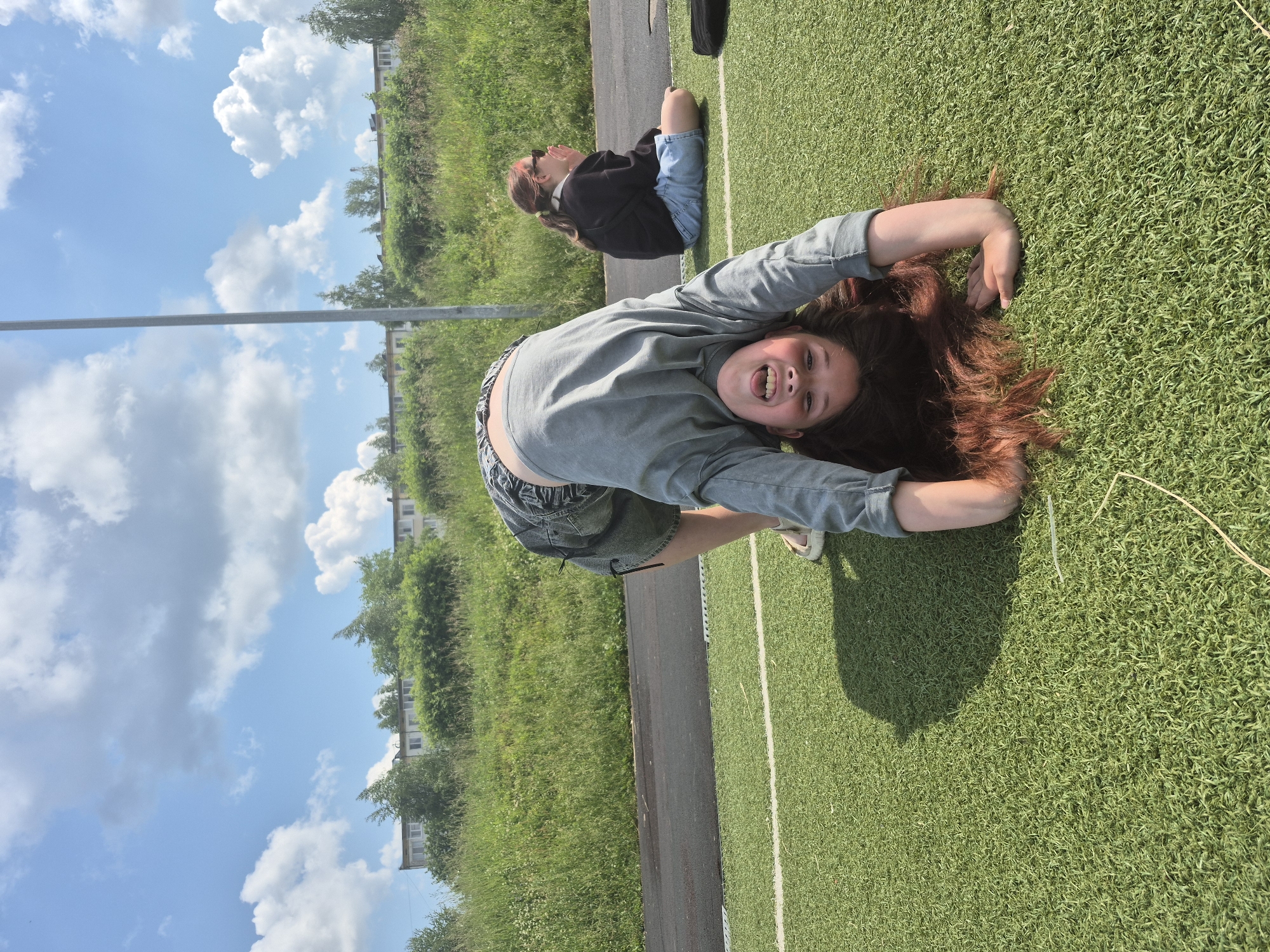 A young girl with long auburn hair is performing a bridge pose on a bright green artificial turf field. She is wearing a gray long-sleeved shirt and denim shorts. Her face is turned towards the camera, smiling with her mouth open, showing her teeth. Her hands are on the ground behind her, supporting her arched body. In the background, to the right, another girl with brown hair tied in a ponytail and wearing a black hoodie and denim shorts is sitting on the turf, facing away from the camera with her hands near her face. Behind them is a dark track, a grassy hill with trees, and a row of apartment buildings. The sky is blue with scattered white clouds, and a tall gray light pole is visible on the right side of the frame. The image was taken outdoors in daylight. The location is the stadium "Progress" in Chaikovsky, Russia.