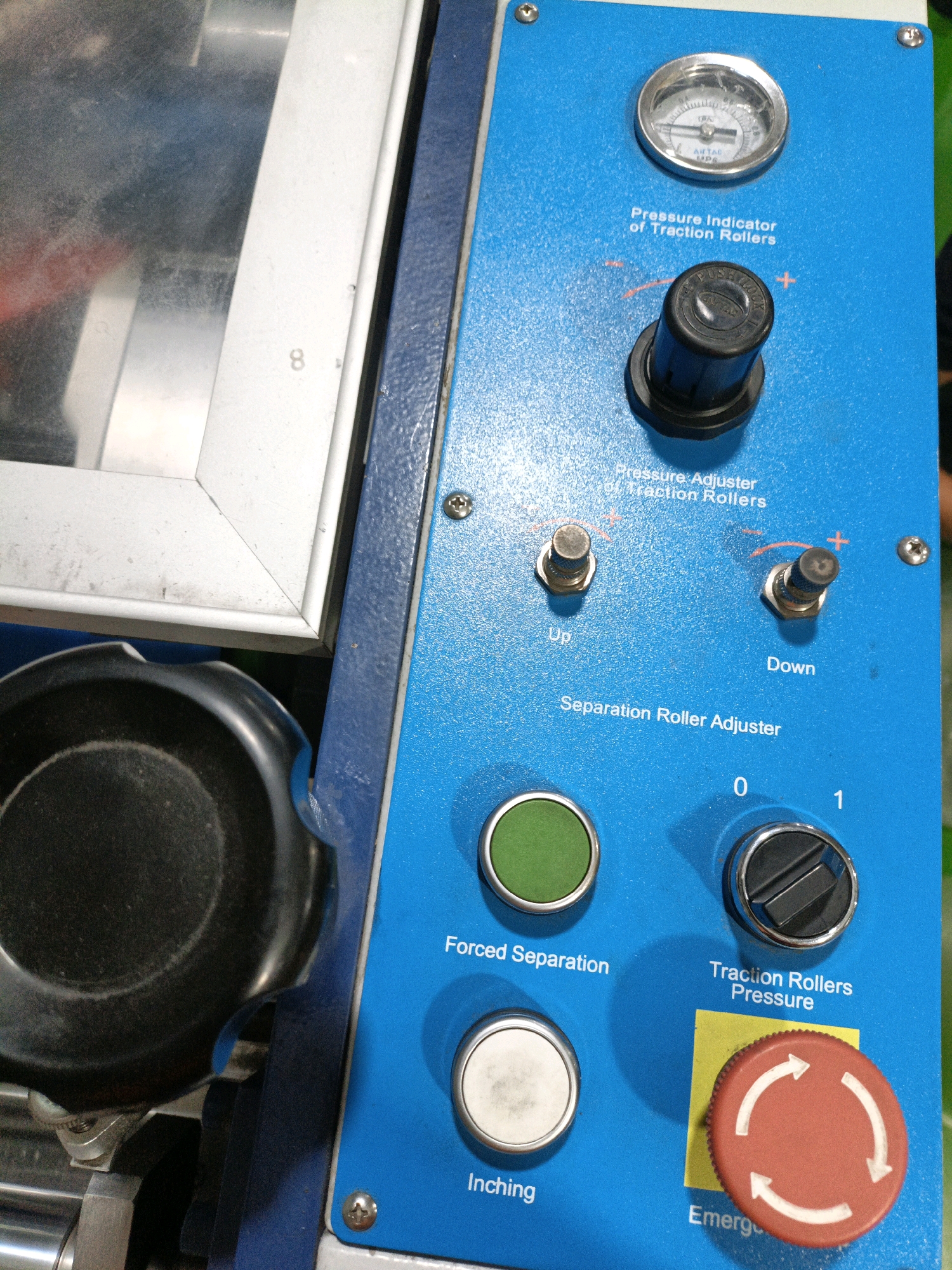 The image shows a close-up view of the control panel of a blue industrial machine. The panel features various dials, buttons, and indicators. Notable elements include a pressure gauge with markings, a black knob labeled "Pressure Adjuster of Traction Rollers" with accompanying "+" and "-" symbols, two toggle switches labeled "Up" and "Down", a green circular button, a white circular button labeled "Inching", a rotary dial with markings "0" and "1" labeled "Traction Rollers Pressure", and a prominent red emergency stop button with circular arrows. The machine appears to be located in an indoor environment, likely a workshop or factory. The background on the left side of the image shows a reflective surface with some white framing, possibly a window or a protective cover. The overall impression is of a functional piece of equipment.