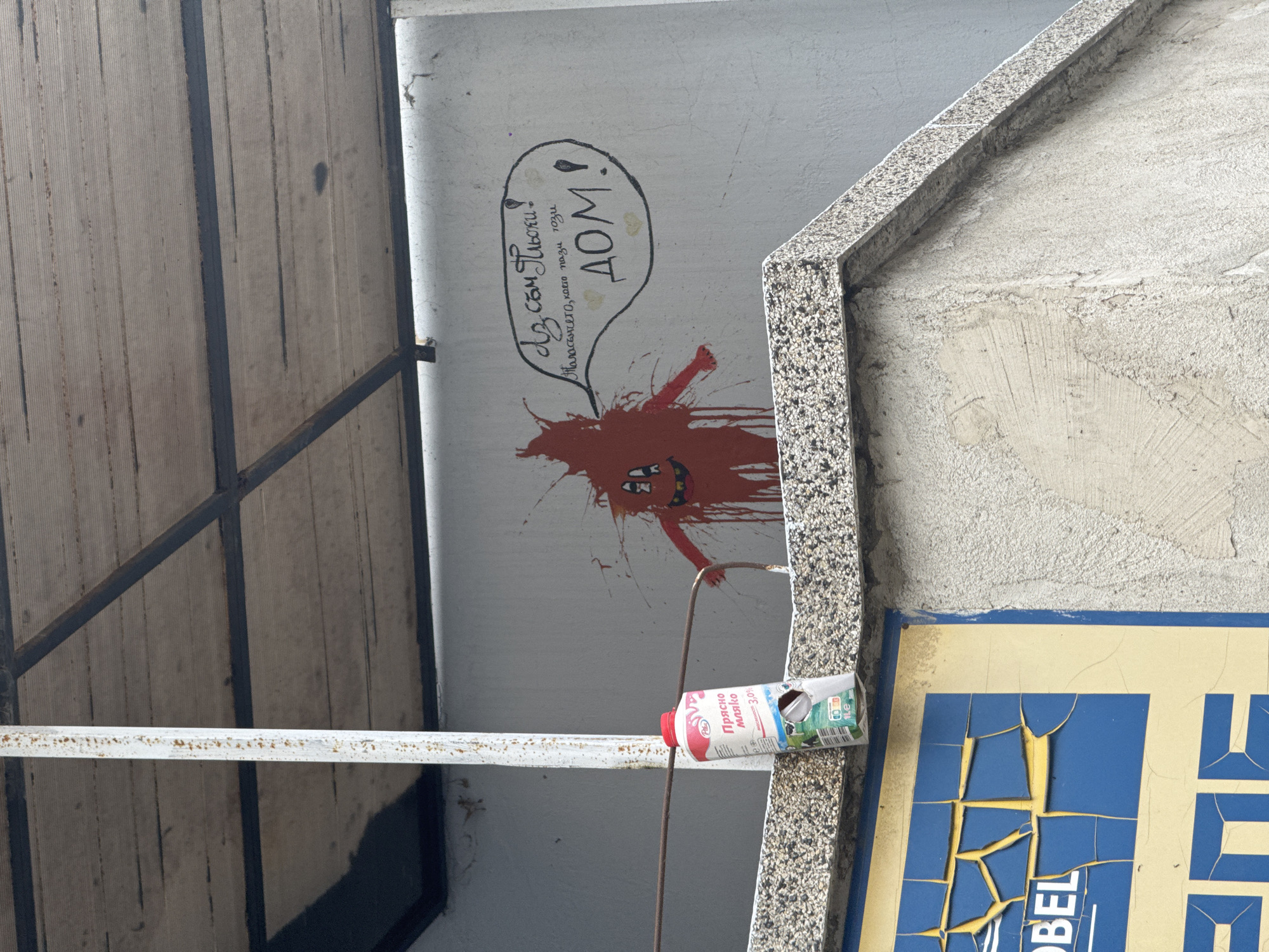 The image shows a section of an urban environment, possibly in Burgas, Bulgaria, based on the Cyrillic writing and the milk carton label. A white wall has graffiti featuring a red, splattered figure with a cartoonish face and the phrase "Аз съм Пльски! ДОМ!! Таласъмчето, което пази този" in a speech bubble. Below the graffiti, a half-crushed milk carton sits atop a white metal railing. To the right is a section of a building with a textured stone edge and a decaying sign with the letters "BEL." A wooden surface with metal supports is visible on the left.
