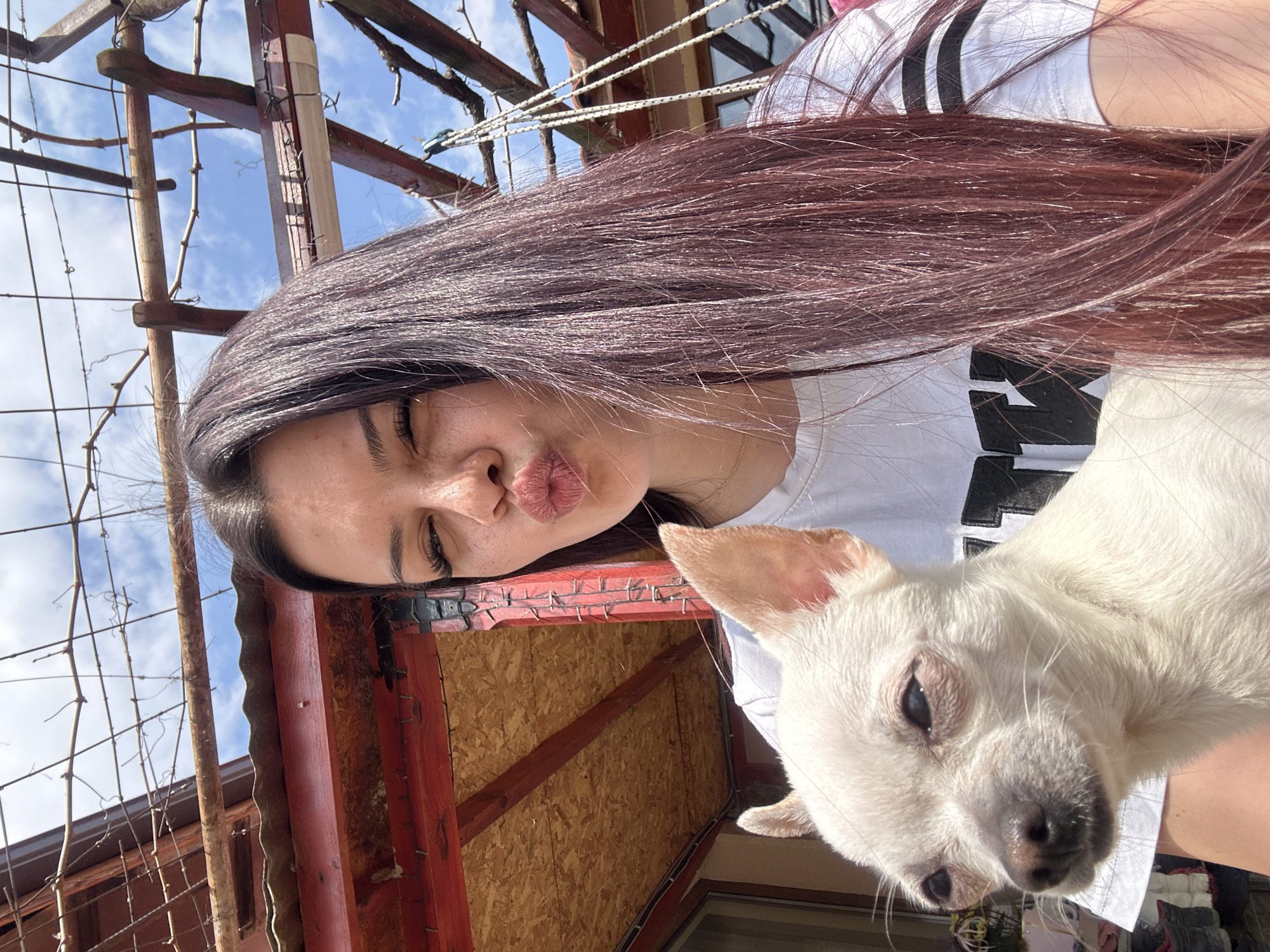 A woman with long, dark, reddish-brown hair is posing for a selfie outdoors with a small white dog. She is puckering her lips in a kissing gesture and wearing a white t-shirt with black stripes. The dog, a chihuahua, is facing forward and appears to be held by the woman. The background includes a wooden structure, possibly part of a pergola or similar outdoor feature, and a glimpse of a blue sky with white clouds. Based on the location information provided, the photo was likely taken in Chubra, Bulgaria, possibly in a garden or outdoor area associated with a residence.