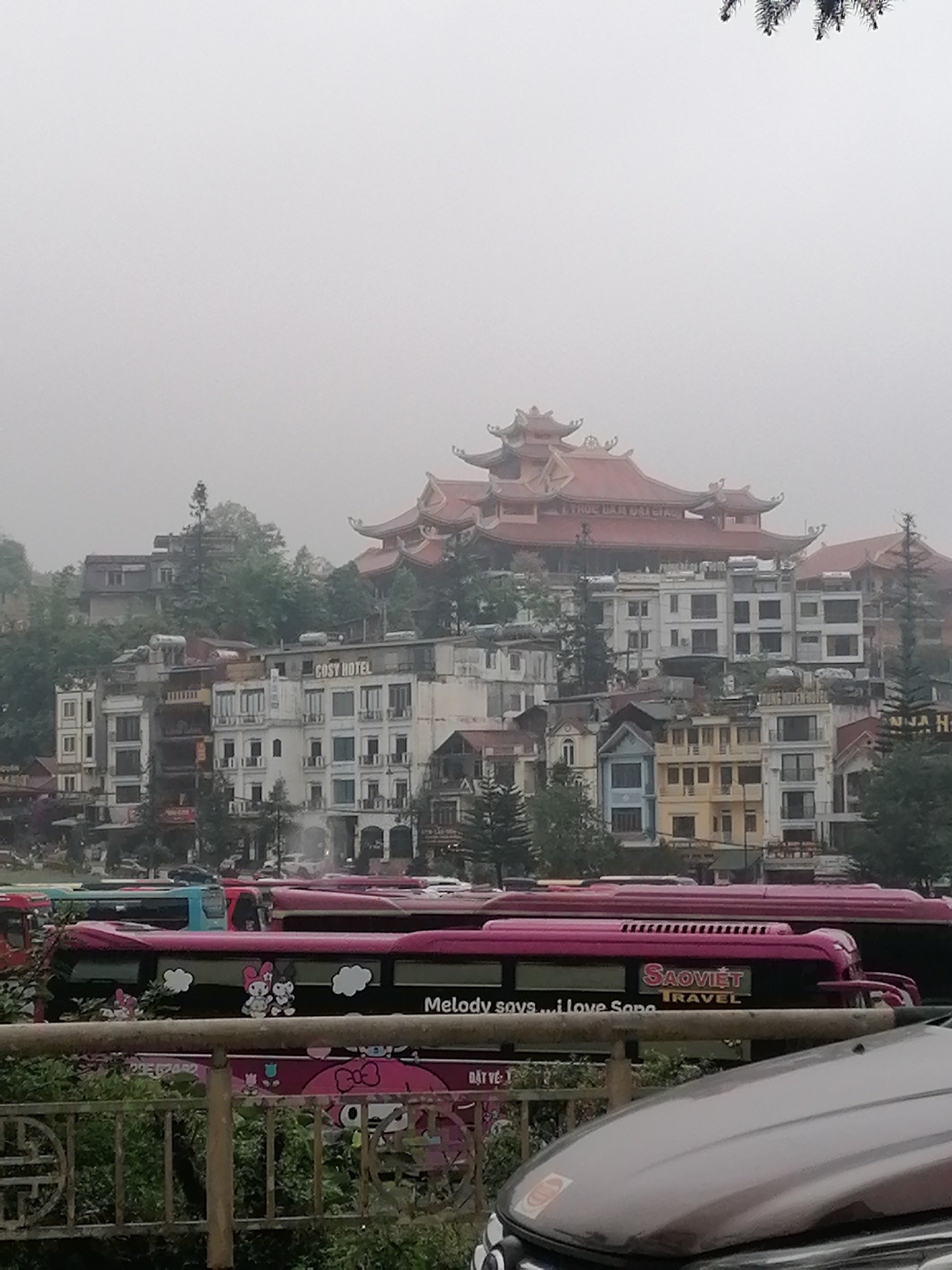 The image shows a foggy, overcast scene in Sa Pa Ward, Vietnam. The background features a tiered building with red roofing in the style of a pagoda, situated above several multi-story buildings, one labelled "COSY HOTEL." In the foreground, there is a row of pink Sao Viet Travel buses with cartoon imagery. A partial view of a vehicle is at the bottom, slightly obstructing the scene. A metal railing and some greenery are also visible.