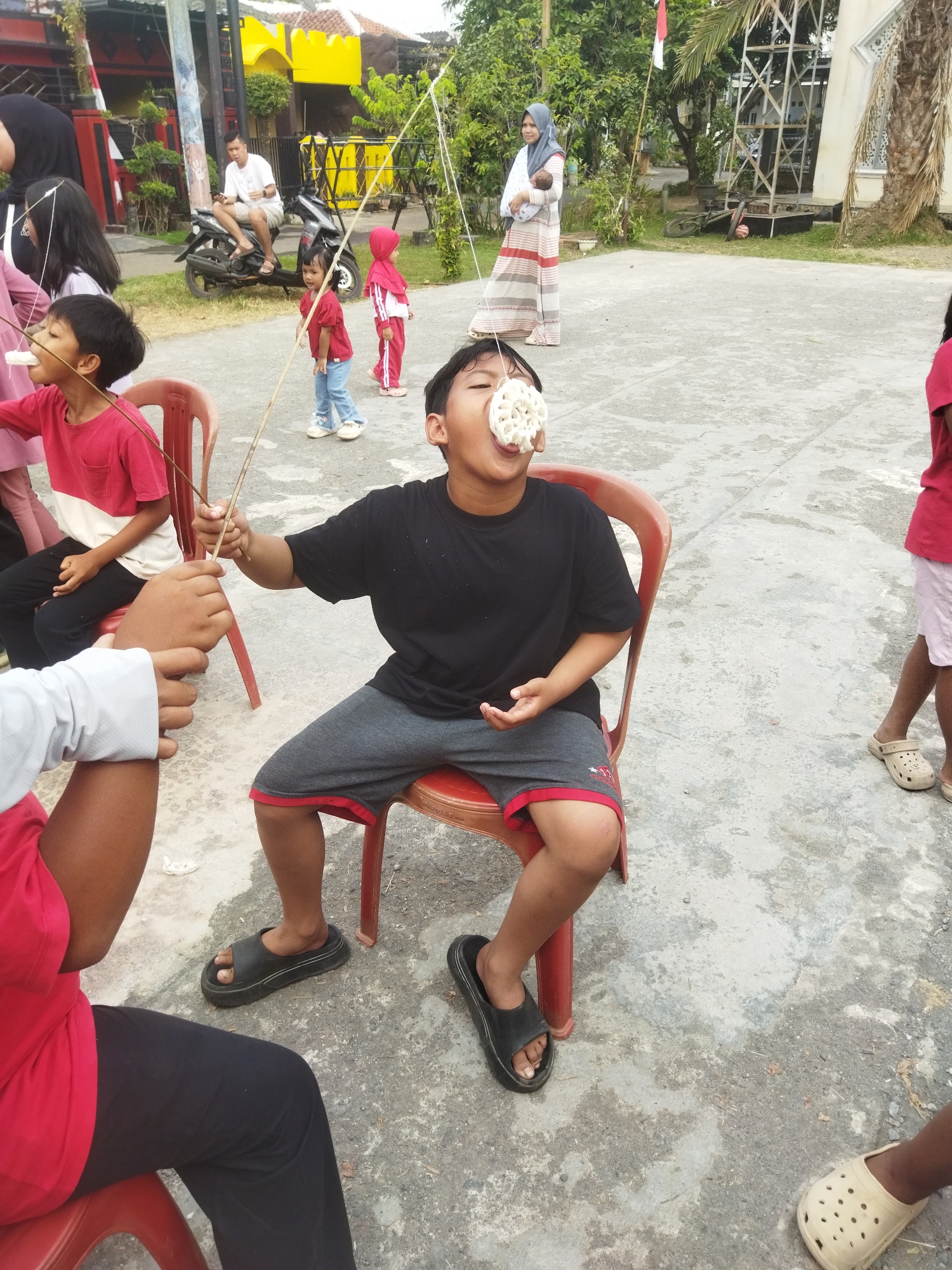 In Banjar, Indonesia, children participate in a cracker-eating contest outdoors. The foreground features a boy in a black t-shirt and gray shorts, seated on a red plastic chair, attempting to eat a cracker suspended by strings. Another child, partially visible in a red shirt and black pants, is seated to his left. In the background, other children, one wearing a red hijab and holding a baby, and another in a red shirt and white pants, are also present. A man sits on a motorcycle behind them. The ground is paved with concrete, and vegetation and buildings are visible in the distance.