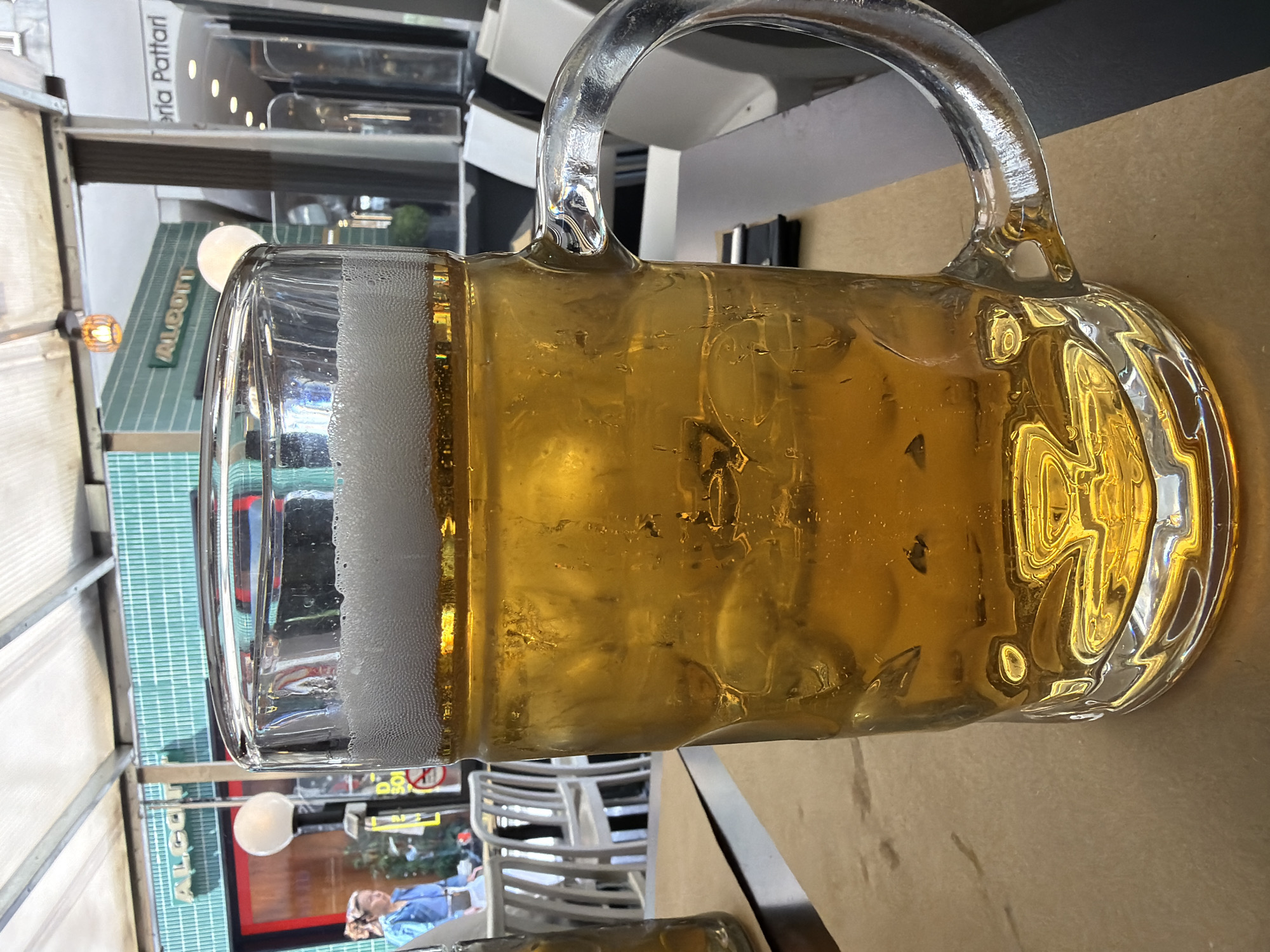 A large glass beer mug filled with beer and ice rests on a tan surface, likely a table. The mug has a thick, clear glass handle and a substantial head of white foam. The beer appears golden in color and contains visible ice cubes. The mug is positioned slightly to the right of the frame, with a tilted perspective.
Outside, a restaurant or bar setting is visible. Through the clear glass of the mug, details of the exterior are distorted but discernible. White plastic chairs are seen in the background, and a person wearing a blue shirt is visible. A sign with the word "ALCOTT" is vertically written in green letters. Another sign, “eria Pattari” is visible above the window.
The photograph was taken in Milan, Italy.