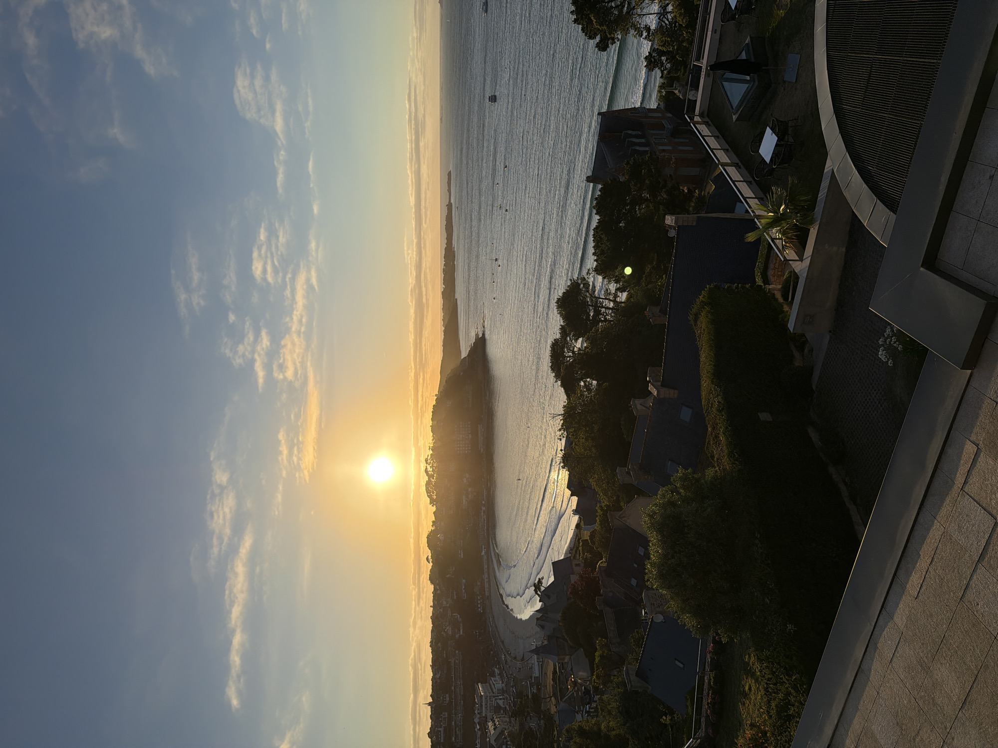 The image depicts a panoramic view of a coastal town during sunset. The sun, a bright orb, is setting over the horizon, casting a warm golden glow across the sky and water. The sky is a gradient of blue and yellow, with scattered clouds tinged with orange.
In the mid-ground, a crescent-shaped beach is visible, with gentle waves lapping at the shore. The water is a deep blue, reflecting the colors of the sunset. Small boats are scattered across the sea. Along the coastline, a town with buildings of varying architectural styles is nestled. The buildings are a mix of traditional and modern, with some featuring red-tiled roofs. Trees and greenery are interspersed throughout the town.
In the foreground, a rooftop terrace is visible, with tables and chairs set up, suggesting an outdoor seating area. The terrace is paved with tiles, and a modern architectural element curves into the frame. This viewpoint overlooks the town and the sea, offering a sweeping vista.
The scene is serene and picturesque, capturing the beauty of a coastal sunset. The location is the commune of La Clarté, located in the Côtes-d'Armor department in Brittany, France. The view is likely from an elevated position, possibly a hotel or a private residence overlooking the bay. No people are visible in the image.