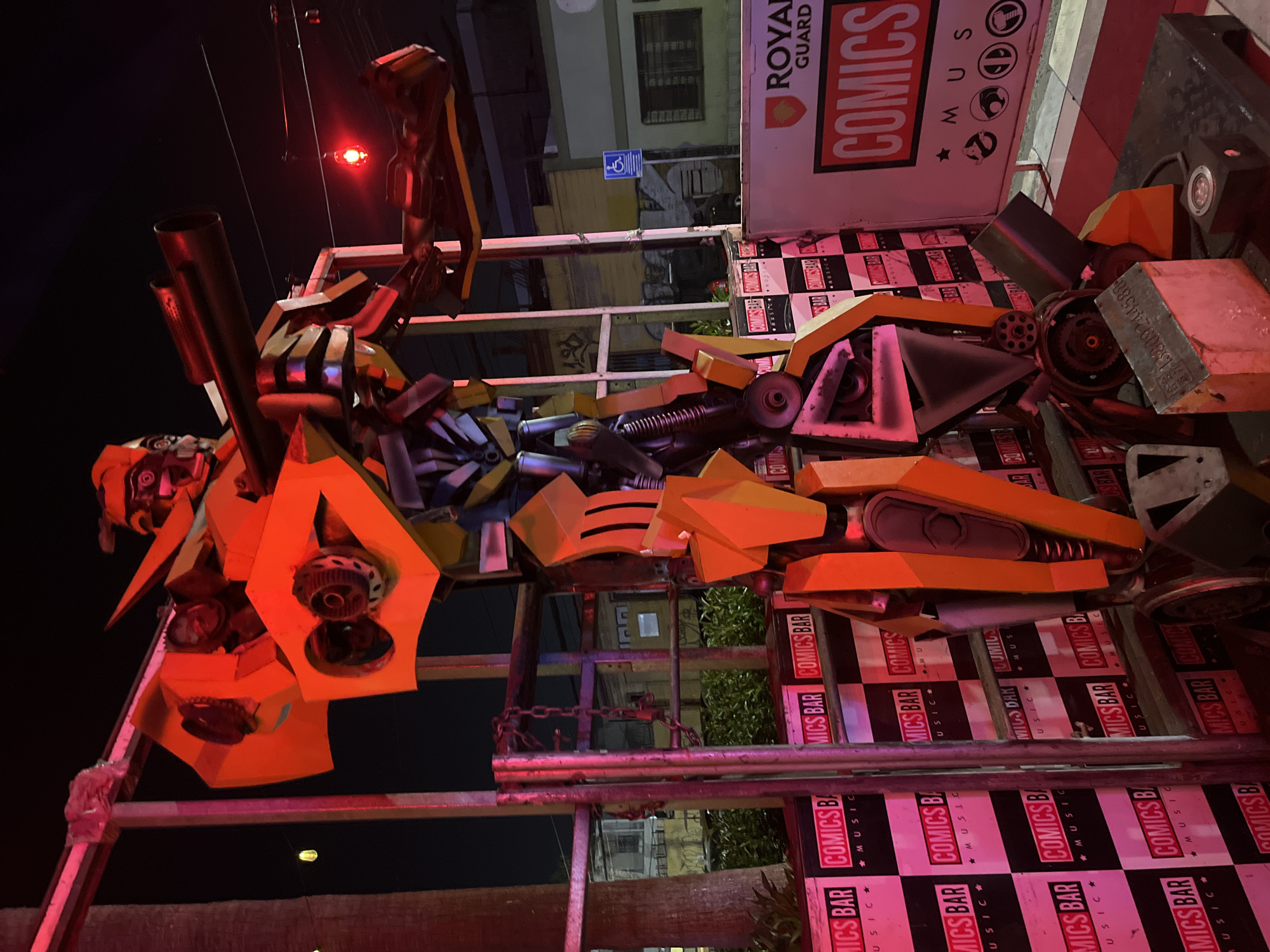 Here is a detailed description of the image:
**General Overview:**
The image presents a fabricated robotic figure resembling a Transformer, likely situated as a decorative piece at the "Comics Bar". The location appears to be urban with a mix of commercial and residential buildings nearby. The shot is taken at night, with artificial lighting illuminating the scene.
**Specific Elements:**
1. **Robotic Figure:** The Transformer-like robot is constructed from various metallic parts and painted in shades of yellow, gray, and blue. It appears to be a static, non-functional sculpture.
2. **Comics Bar:**
* The bar's name and logo ("Comics Bar") are repeatedly printed on the background panels, predominantly in red and white colors.
3. **Background:**
* Elements of the urban environment are visible, including portions of buildings.
* Signs, including one with accessibility symbols.
4. **Lighting:** The primary lighting source is a red light situated in the background. Additional artificial lights illuminate the bar and robot, creating a warm, saturated tone.
5. **Environment:**
* The figure is supported by metal scaffolding, likely erected to hold the heavy structure.
* Plants are seen surrounding the location.
**Actions and Interactions:**
None of the figures are performing any actions.
**Location:**
Based on the OCR of the bar name "Comics Bar" I can't provide any location for the image. The city and country are unknown.
**Overall Impression:**
The image captures an unusual urban scene, featuring a creative installation that combines robot aesthetics with the branding of a local bar.