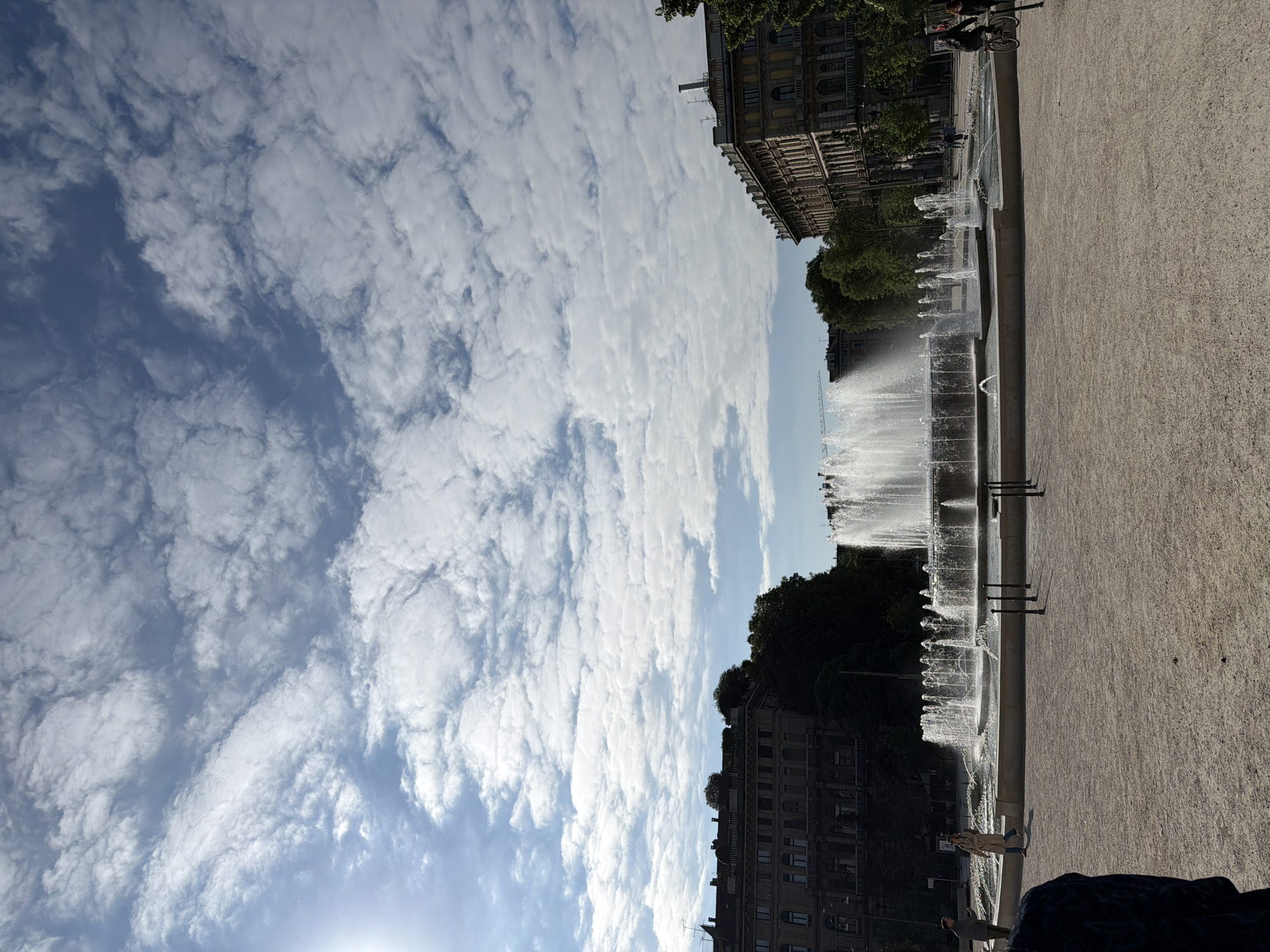This media file presents an urban square in Milan, Italy, featuring a long, modern fountain under a bright, partly cloudy sky. The central subject is a multi-tiered fountain spanning horizontally across the mid-ground, where numerous jets of water spray upwards, creating an active and dynamic display.
On the left and right sides of the square, behind the fountain, are several traditional European buildings. These multi-story structures feature ornate facades, numerous windows, and some balconies, indicative of typical Milanese architecture. Green trees are interspersed along the building lines, adding natural elements to the urban landscape.
The foreground is dominated by a wide expanse of light-colored, textured pavement, likely stone or concrete, suggesting a public plaza or pedestrian area. Several people are visible within the scene: one individual is seen on a bicycle in the upper right portion of the square, while another person wearing a light-colored coat walks alongside the fountain in the lower right. Other figures can be vaguely seen in the distance, suggesting a moderately busy public space.
The sky is a mix of clear blue patches and scattered, fluffy white clouds, indicating fair weather. The bright illumination suggests it is daytime, likely morning or early afternoon. The overall atmosphere conveyed is one of urban leisure and pleasant activity. There is no discernible text visible in the image.