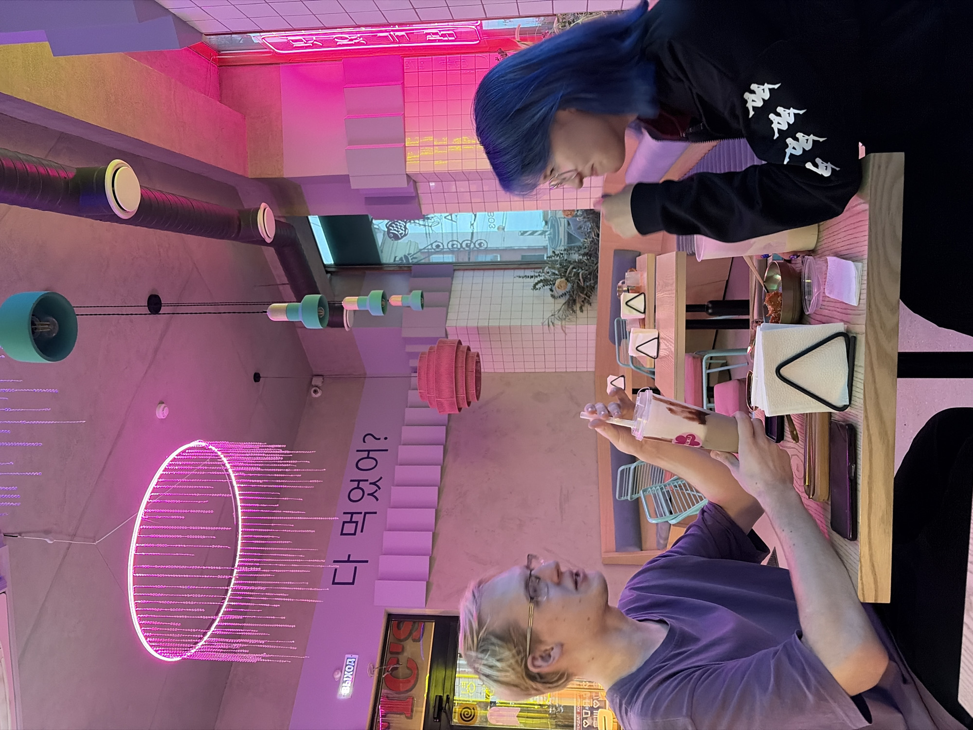 This image depicts two individuals seated at a light wooden table inside "TIC'S Korean Chicken & Drinks," located at Pervomayskaya Street, 208, Izhevsk, Russia.
A male with short blonde hair and glasses, wearing a purple t-shirt and dark trousers, is seated on the left. He holds a clear plastic cup containing a layered light brown and white beverage with a straw in his right hand. His mouth is slightly open, indicating he is speaking. Opposite him, a female with long, bright blue hair and glasses is seated. She wears a black long-sleeved jacket adorned with multiple white Kappa logos on her left sleeve. Her right hand is positioned near her chin as she looks towards the male.
On the table between them, a white triangular napkin dispenser, a dark smartphone, and a small metallic bowl containing a portion of reddish food are present. A second clear plastic cup with a light-colored beverage is partially visible on the female's side of the table, along with a small transparent container holding a white square object.
The cafe interior features a concrete ceiling with exposed dark gray ventilation ducts. Several teal-colored pendant lights hang at varying heights. A large, circular pink neon chandelier with numerous hanging white decorative strands is prominent in the upper left. The walls are light pink or purple, with Korean script "다 먹었어?" (Did you eat it all?) displayed prominently. A series of horizontal rectangular panels are mounted below the Korean text. A white sign with Russian text "ВЫХОД" (Exit) is illuminated with a purple glow above a doorway on the far left. White tiled walls with bright pink neon strip lighting are visible on the right side of the room. Booth seating with light blue upholstery and wooden backs, along with chairs featuring teal frames, are visible in the background. The overall environment is brightly lit with a dominant magenta and cyan light scheme.