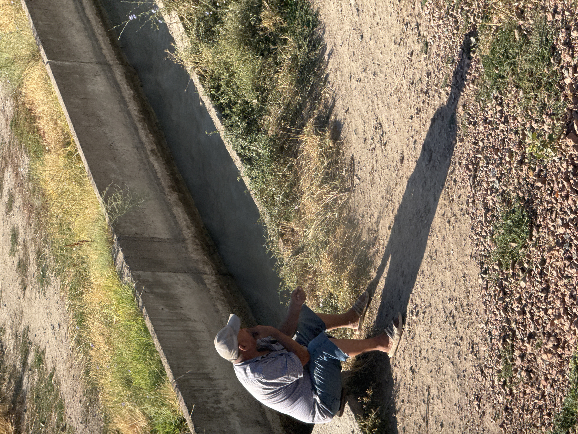 A man wearing a light-colored baseball cap, a checkered short-sleeved shirt, and blue shorts sits on a concrete ledge beside an irrigation canal. He is on the right side of the frame, his body angled away from the camera and towards the canal. His right hand is near his face, and his left hand is extended slightly, holding what appears to be a small object, possibly a cigarette or a piece of straw. He is wearing light-colored sandals. The irrigation canal is filled with dark water. The ground is dirt and gravel, with patches of dry and green grass growing along the sides of the canal and on the bank where the man is sitting. The sun casts a long shadow from the man onto the ground. The image was taken in Ivanovka, Kyrgyzstan.