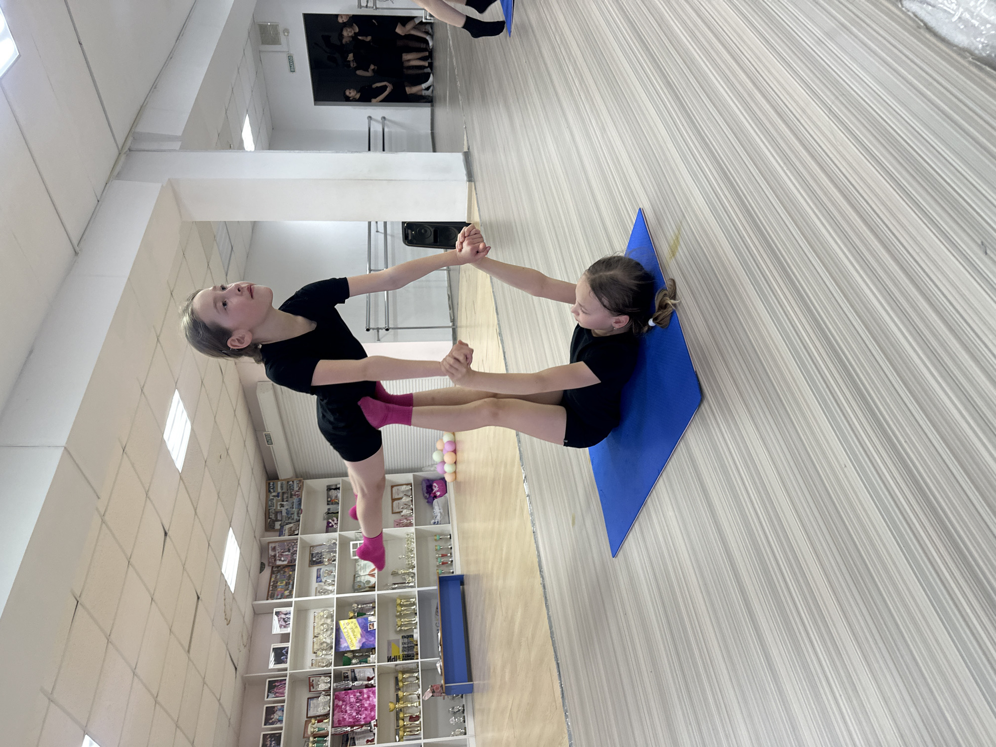 Two young girls, likely pre-teens or early teens, are captured mid-pose in an indoor gymnasium or dance studio in Votkinsk, Russia. Both are dressed in black athletic attire; one girl wears bright pink socks. They are engaged in a partner acrobatic or gymnastic exercise, demonstrating flexibility and balance. One girl lies on her back on a blue mat, with her legs raised towards the ceiling. She is holding the hands of the second girl, who stands on one leg, balancing on her toes, with her other leg raised. Their arms are extended, forming a symmetrical "V" shape. Both girls appear focused on their pose.
The setting is a brightly lit room with white walls and a light-colored, striped laminate floor. Rectangular ceiling lights illuminate the space. In the background, a large white shelving unit is filled with numerous trophies and awards, suggesting a history of achievements in sports or performance. A large mirror on one wall reflects blurred figures of other individuals, indicating the presence of a class or practice session. A black sign on the wall clearly displays the Russian word "ВЫХОД," meaning "EXIT." The scene suggests a daytime practice session under artificial lighting.