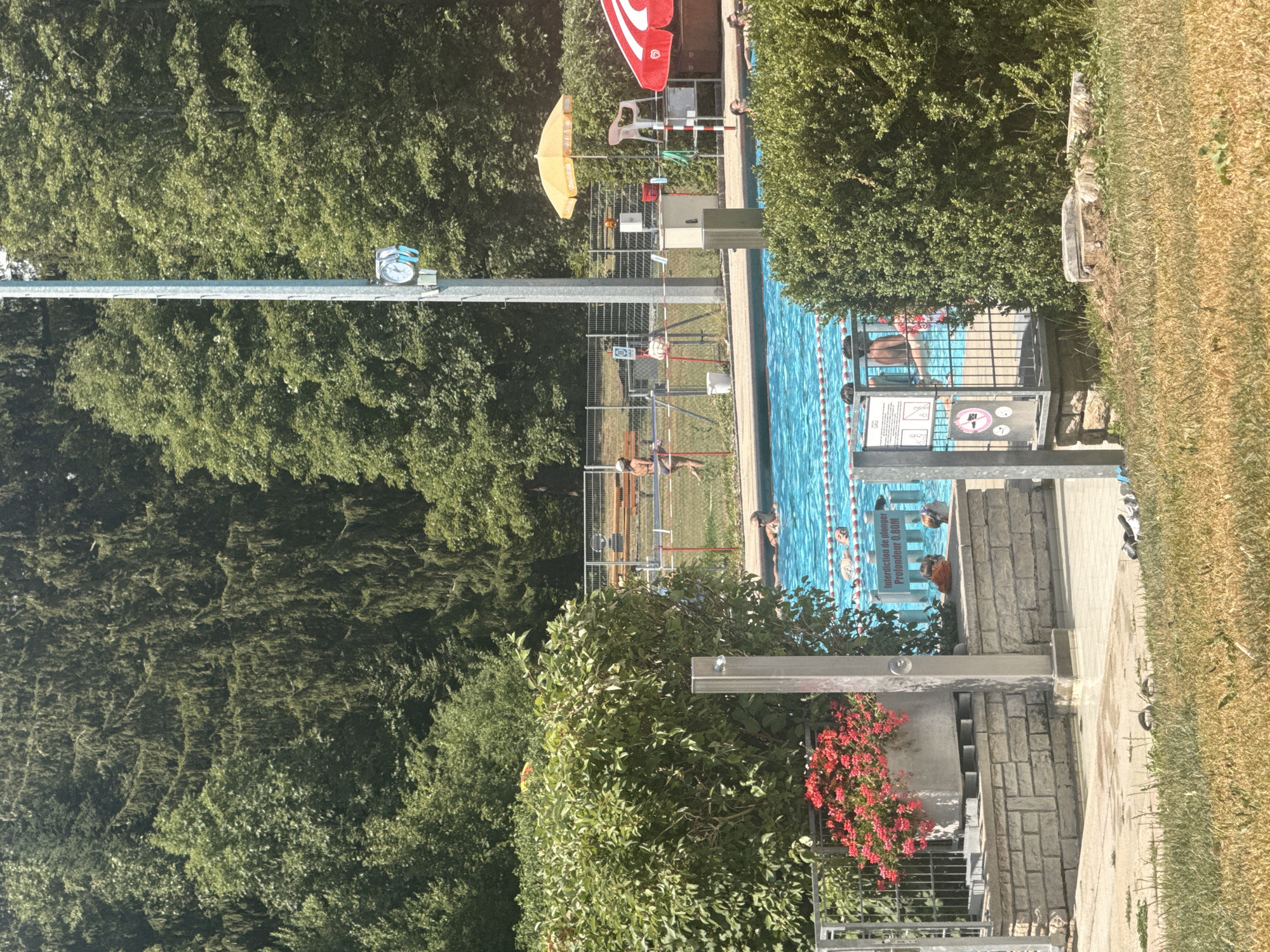 The image depicts a public outdoor swimming pool in Le Locle, Switzerland. The pool is filled with blue water, and several people are visible enjoying it. Two individuals are partially submerged, appearing to swim or float. In the background, on the pool deck, a lifeguard chair with a red and white striped umbrella is present, along with a yellow umbrella. A tall metal pole with a clock attached to it stands near the pool. Lush green trees and foliage surround the swimming area, suggesting a park or natural setting. The pool area is bordered by metal fencing and stone walls, with some decorative flowering plants in a planter. A sign in French indicates a diving prohibition with a depth of 0.80M. The overall scene suggests a warm, sunny day at a recreational facility.