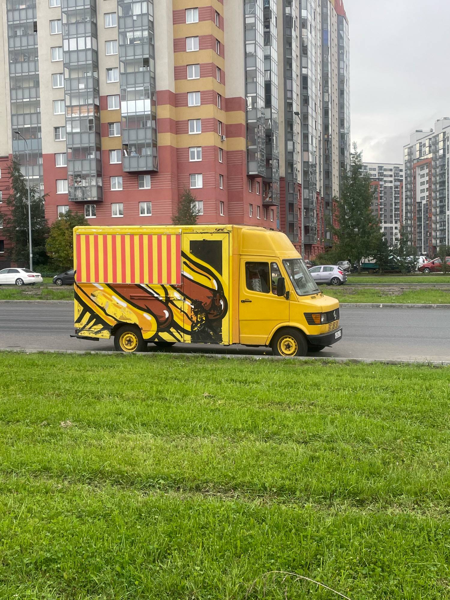 A yellow food truck with a graphic of a hot dog on its side is parked on the side of a road in front of a residential building in Saint Petersburg, Russia. The food truck has a yellow and red striped awning. The building is tall and modern, with multiple balconies. A grassy area is in the foreground.