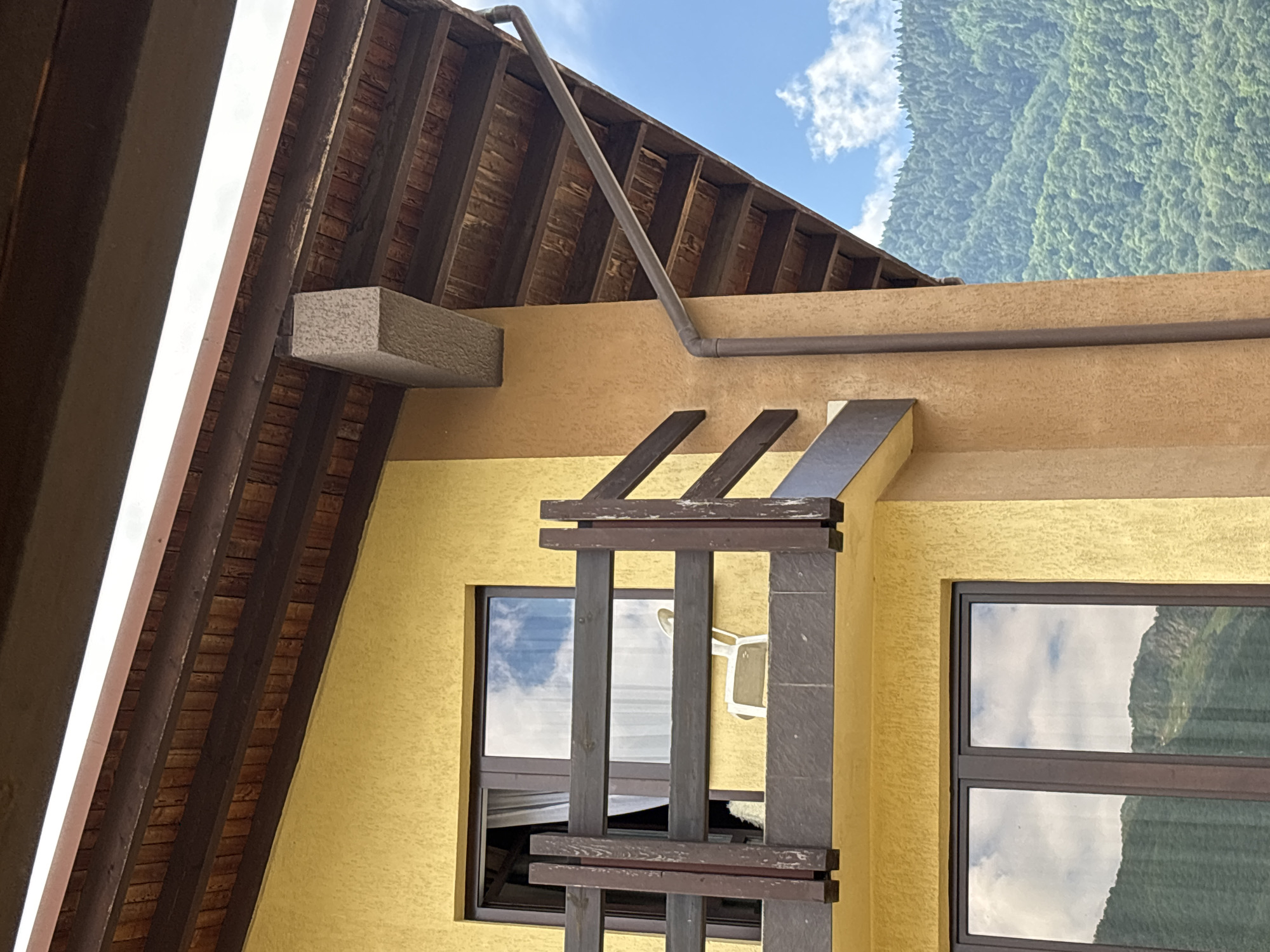The image shows the exterior of a building in Krasnaya Polyana, Russia, featuring a yellow-colored textured facade with dark brown wooden window frames and a balcony railing. The balcony has a white plastic chair visible. Above the balcony, the exposed wooden beams of a roof structure are visible, along with a brown downspout. In the background, a lush green mountainside covered in trees rises against a blue sky with scattered white clouds. The lighting suggests it is daytime.