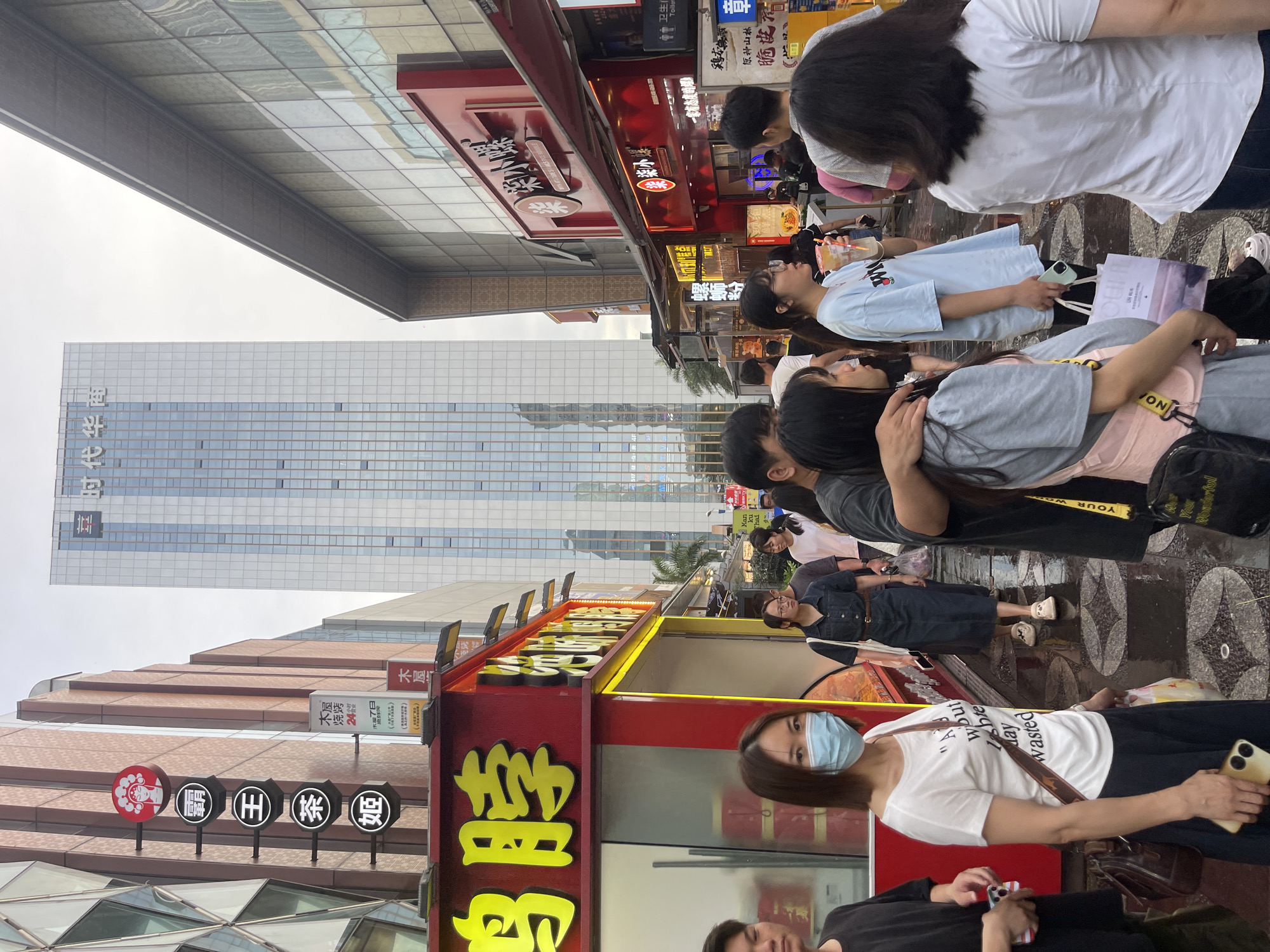 The image depicts a street scene in Panyu District, China, with a focus on a bustling pedestrian area lined with shops and food stalls. A tall, modern glass-fronted building dominates the background, reflecting the sky and surrounding structures. In the foreground, a diverse group of people are walking along a wet pavement, suggesting recent rain. Many are dressed casually, and some are carrying shopping bags.
On the left side of the street, a red and yellow food stall features prominent Chinese characters, indicating it is a place selling food. Several circular signs with Chinese text and imagery, possibly representing a brand or type of cuisine, are mounted vertically on a building to the left of the food stall. A woman in a white t-shirt and a light blue face mask is walking towards the viewer, holding a phone and a shopping bag.
Further down the street, more shops and stalls are visible, also displaying Chinese signage. The architecture suggests a commercial district. The overall atmosphere is one of everyday urban life, with people engaged in shopping, dining, or simply navigating the street. The wet ground and overcast sky contribute to a slightly subdued lighting condition.