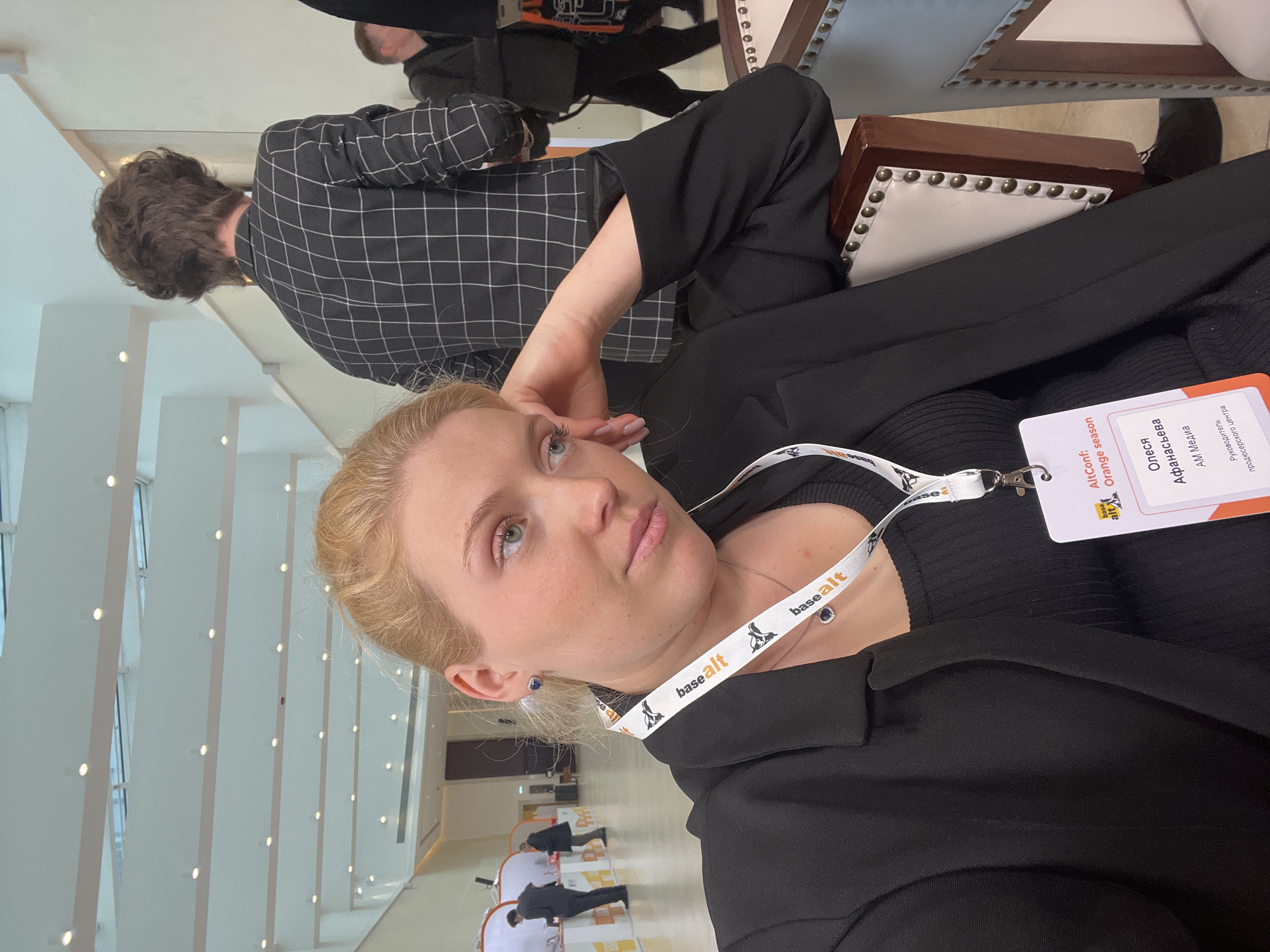 The image shows a woman, possibly named Olesya Afanasyeva from AM Media, wearing a black blazer and a lanyard with a "basealt" logo and a name tag. The tag indicates that she is at "AltConf: Orange season." She's sitting on a chair with her hand near her face. Behind her is a person in a black and white checkered shirt. The location appears to be an interior space with white walls and a row of windows. Further in the background are other people standing or seated. Based on the name on the conference tag, it's possible the location is connected to BaseALT, a Russian software company.
Moscow, Russia is the home of BaseALT.