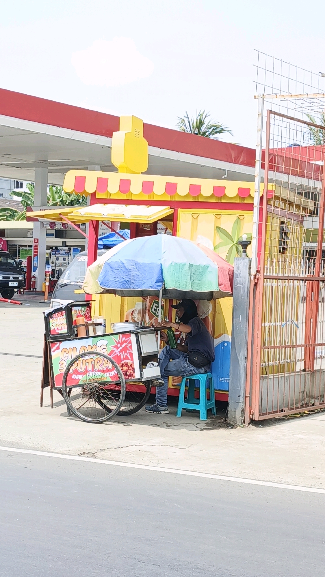 The image depicts a street vendor in Desa Wonoharjo, Indonesia, operating a food cart under a colorful umbrella. The vendor, wearing a dark t-shirt, blue jeans, and a black cap, is seated on a blue stool to the right of the cart, holding a green bottle. The food cart, adorned with a sign that reads "CILOK 3 PUTRA," displays various food items. Behind the cart is a yellow kiosk with red and yellow awnings, and further in the background, a red and white gas station canopy is visible, suggesting the location is near a road. A metal gate stands to the right of the scene, with a palm tree visible in the distance. The overall environment appears to be an outdoor, urban setting.