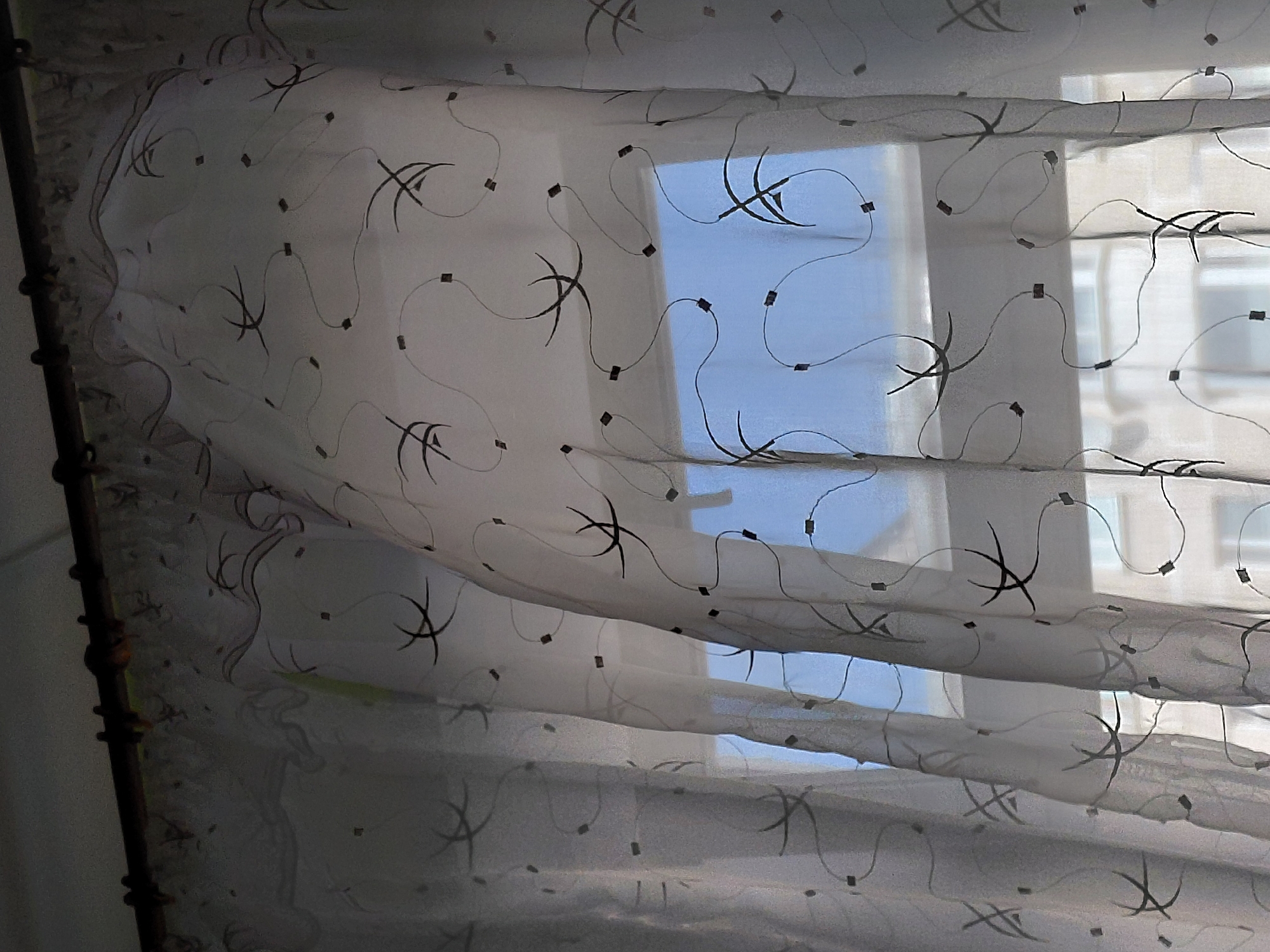 The image captures an indoor view looking outwards through a window in Nadym, Russia. The main subject is a sheer, translucent white curtain, intricately patterned with stylized black squiggly lines and small black square accents. The fabric appears voluminous and slightly billowed, suggesting either a gentle breeze or its natural draped state.
Beyond the curtain, a window frame is visible, through which a bright, clear blue sky is discernible. Parts of light-colored buildings with multiple windows can be seen in the distance, indicating an urban or suburban environment. On the far left, a dark curtain rod with a series of rounded, decorative elements is partially visible.
The scene is set during the daytime, evidenced by the bright light and blue sky, implying fair weather. There are no people present, and no specific activity is taking place beyond the potential subtle movement of the curtain. No visible text is present in the media file. The image offers a tranquil glimpse from an interior space to the exterior world.