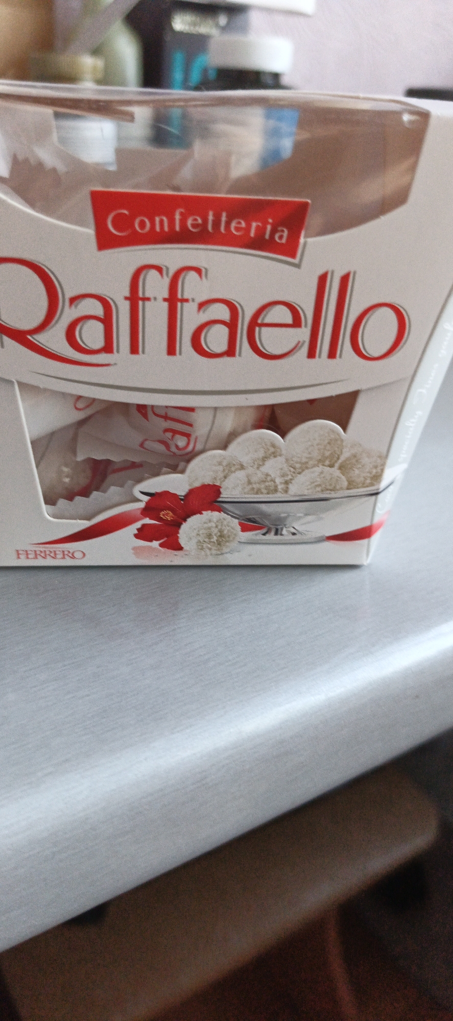 The image shows a close-up of a box of RaffaHELLO chocolates. The box is white with red lettering. The words "Confetteria" and "RaffaHELLO" are prominently displayed. Below the brand name, there are several RaffaHELLO chocolates visible, some in their wrappers and some unwrapped, sitting in a clear plastic container. There is also a red ribbon and a red flower accent on the front of the box. The box is resting on a light grey surface, which appears to be a table or countertop. The background is blurred, showing some other objects, possibly other products or packaging. There are no people in the image. The scene is indoors, and based on the provided information, it is located in the city of Tikhvin, Russia.