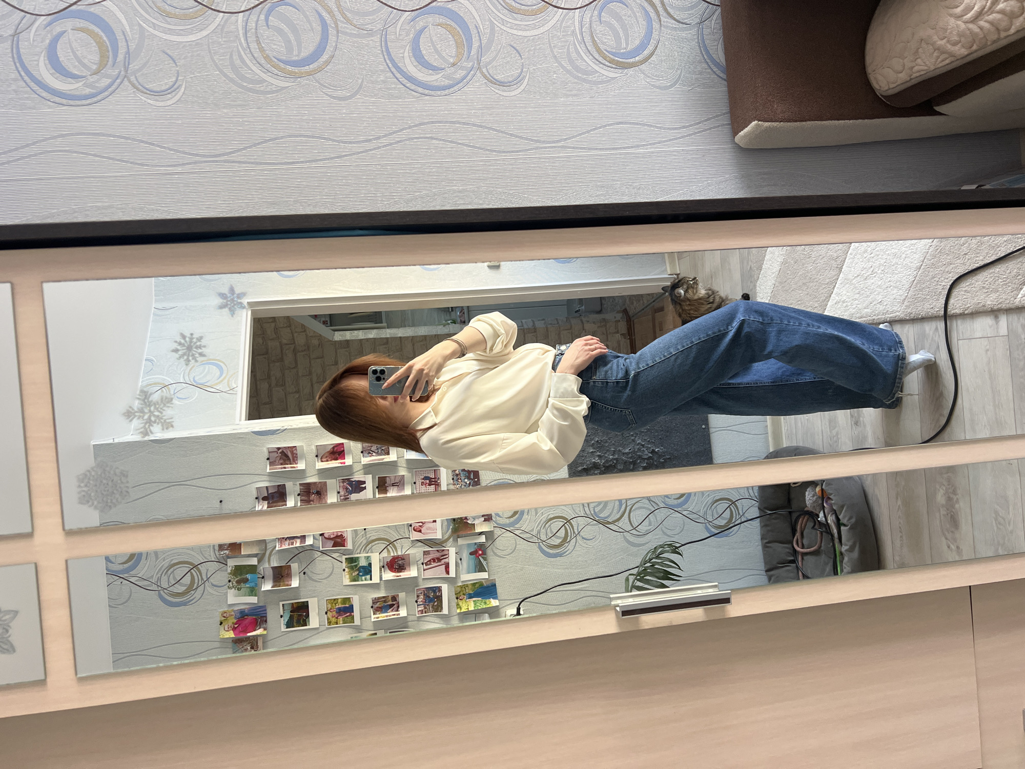 A person with reddish-brown hair, wearing a cream-colored collared shirt and wide-leg blue jeans, stands facing a mirror, taking a selfie with a grey smartphone. The individual's face is obscured by the phone. A silver bracelet is visible on the right wrist. The mirror is part of a light wood-framed wardrobe or cabinet. Attached to the mirror are numerous small, rectangular photographs.
Reflected in the mirror is a room with light blue and grey wallpaper featuring abstract circular and wavy patterns. Snowflake decals are present on some mirrored and wall surfaces. On the light-colored wooden floor, a black power cord runs along the ground. A fluffy, brown and black striped cat is visible near the person's feet, looking to the right. A dark grey pet bed containing a rope toy and other items is reflected further back on the floor. Behind the pet bed, a large green potted plant is partially visible. To the right of the reflection, a brown upholstered furniture piece with a light-colored, patterned cushion is present. A doorway with a light-colored frame leads to another area with a brick-patterned wall.
This scene is located within a residential interior in Chaikovsky, Russia.