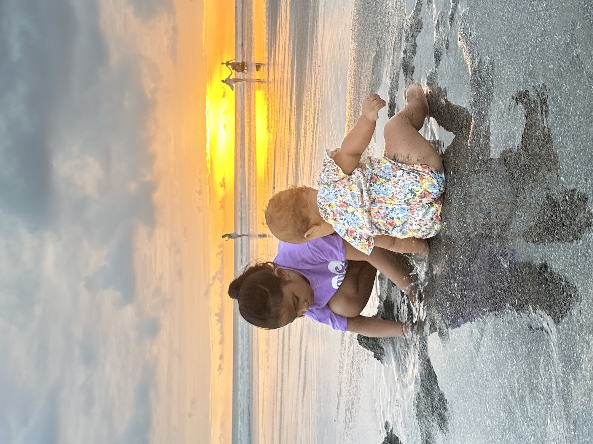 In North Kuta, Indonesia, two young children play on a beach at sunset. The child on the left, wearing a purple t-shirt, squats in shallow water, digging in the sand. The child on the right, in a floral romper, sits facing away from the camera, also interacting with the sand and water. The ocean stretches out behind them, reflecting the vibrant orange and yellow hues of the setting sun. The sky is filled with dramatic clouds, creating a warm and atmospheric backdrop. In the distance, silhouetted figures can be seen walking along the shoreline. The wet sand glistens, and the shallow water creates reflections of the children and the sky.