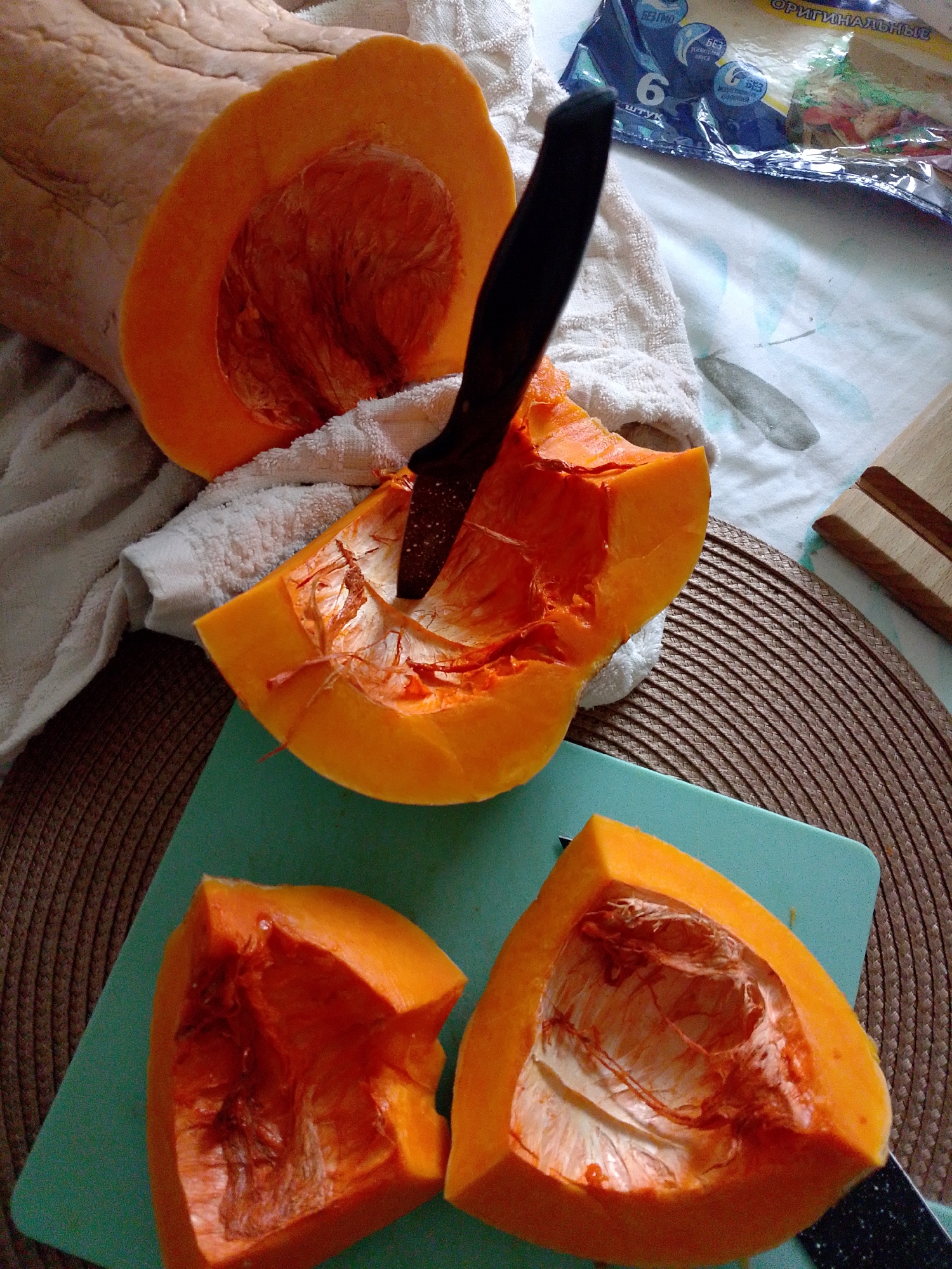 The image shows a cut butternut squash on a green cutting board on a brown woven mat. The squash is cut into four pieces, with a black-handled knife stuck into one piece. A white towel is visible behind the squash and cutting board. A package of tortillas is visible in the background, as is a wooden stand. The location is Izhevsk, Russia, and the image appears to be taken indoors.