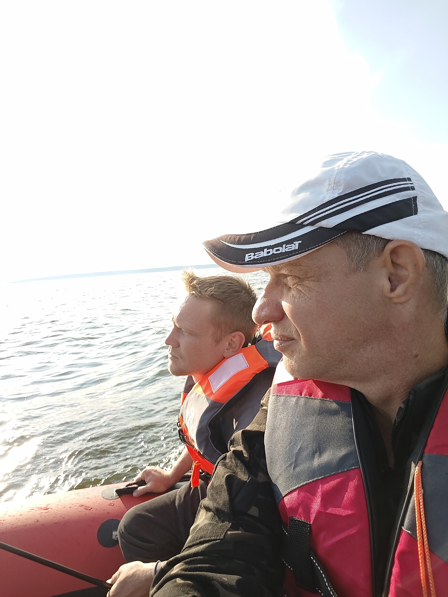 Two men are in a red inflatable boat on the Kama River near Chaykovsky, Russia. The man in the foreground is wearing a white baseball cap with black stripes and the logo "Babolat." He is also wearing a red and grey life vest. The man in the background is wearing an orange and grey life vest. The water is rippled, and the sky is bright and overcast. The men are looking out at the water.