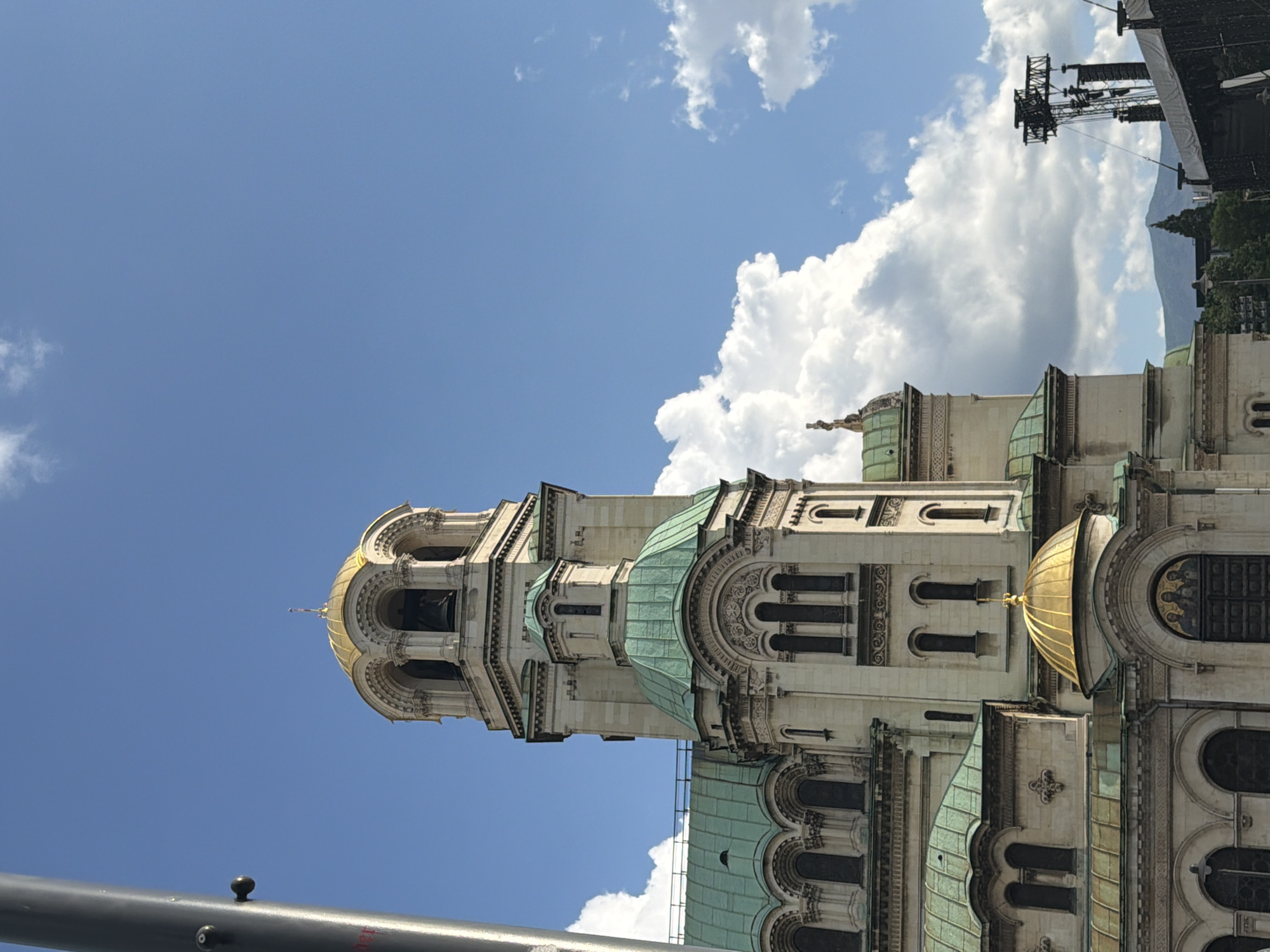 The image displays a detailed view of the St. Alexander Nevsky Cathedral in Sofia, Bulgaria, under a clear blue sky with white cumulus clouds, primarily clustered on the right side. The cathedral's facade is constructed from light-colored stone, featuring multiple green-patinated copper domes and roofs, alongside two prominent gold-leafed domes. The main bell tower, located on the right side of the cathedral, is tall and intricate, culminating in a large golden dome with a spire, and features arched openings that house bells. A smaller golden dome is visible lower down on the facade, above an ornate archway containing a mosaic depicting three figures. Arched windows and decorative stone carvings are visible throughout the building. A thick, dark grey cylindrical pole occupies the far left of the frame, partially obscuring the cathedral and featuring a black, knob-like protrusion and faint red lettering near its base. In the background on the far right, a large black metal scaffolding structure, possibly for an event stage, with attached speakers and a white tarpaulin covering, is visible. Beyond this structure, a hazy mountain range and green trees are discernible against the horizon. No people are present in the scene.
