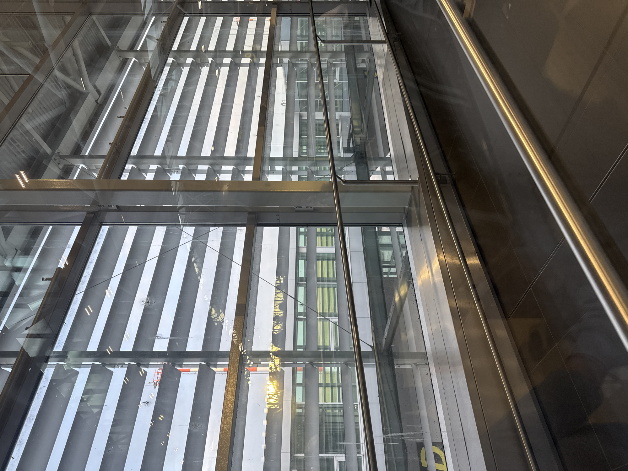 The image shows an interior perspective through a glass wall, potentially in a building in Bratislava, Slovakia. Reflections on the glass indicate another building with vertical striped patterns in gray, white, and some other colors. The frame of the glass wall is metal with a reflective surface, and a metallic pole runs vertically through the image. The scene seems to be taken from inside a building, and the focus is on the exterior environment as viewed through the glass structure.