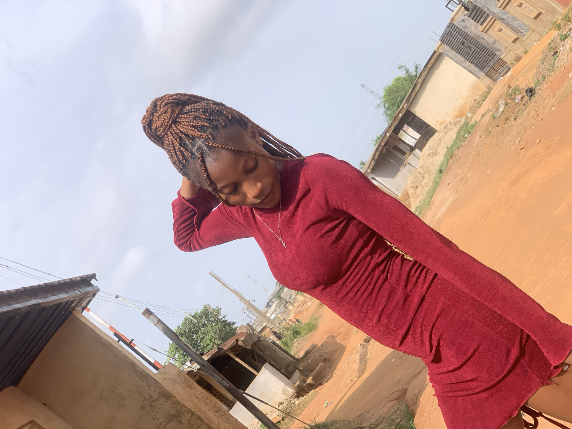 A young woman stands outdoors in Ikorodu, Nigeria. She is wearing a long-sleeved, maroon dress and has her braided hair styled in an updo. Her head is tilted down, and her hand rests on her head. A silver cross necklace is visible on her chest. The background consists of a dirt road, simple buildings constructed of concrete and corrugated metal, and overhead power lines. The sky is overcast.