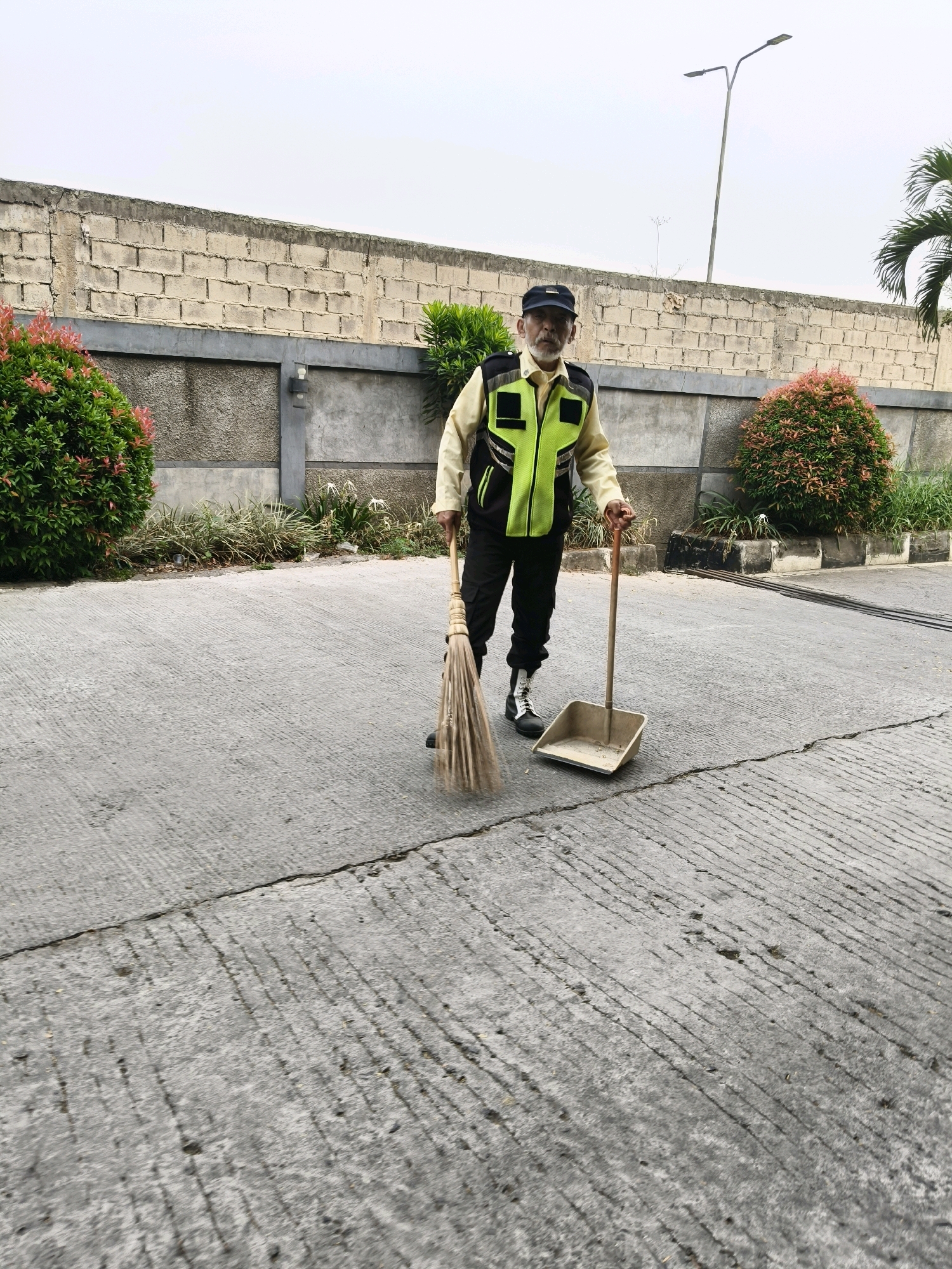 An elderly man wearing a yellow shirt, black pants, a high-visibility lime green vest with black accents, and a dark blue cap is sweeping the ground with a broom and dustpan. He has a gray beard and mustache. He is standing on a concrete surface with visible lines and cracks. Behind him is a textured gray wall, a line of plants with white flowers, and two bushes with pink and green leaves. A tall streetlight is visible in the background. The scene appears to be outdoors, possibly in a public area or private property. The lighting suggests it is daytime. No specific location in Bandung City, Indonesia is evident from the image.