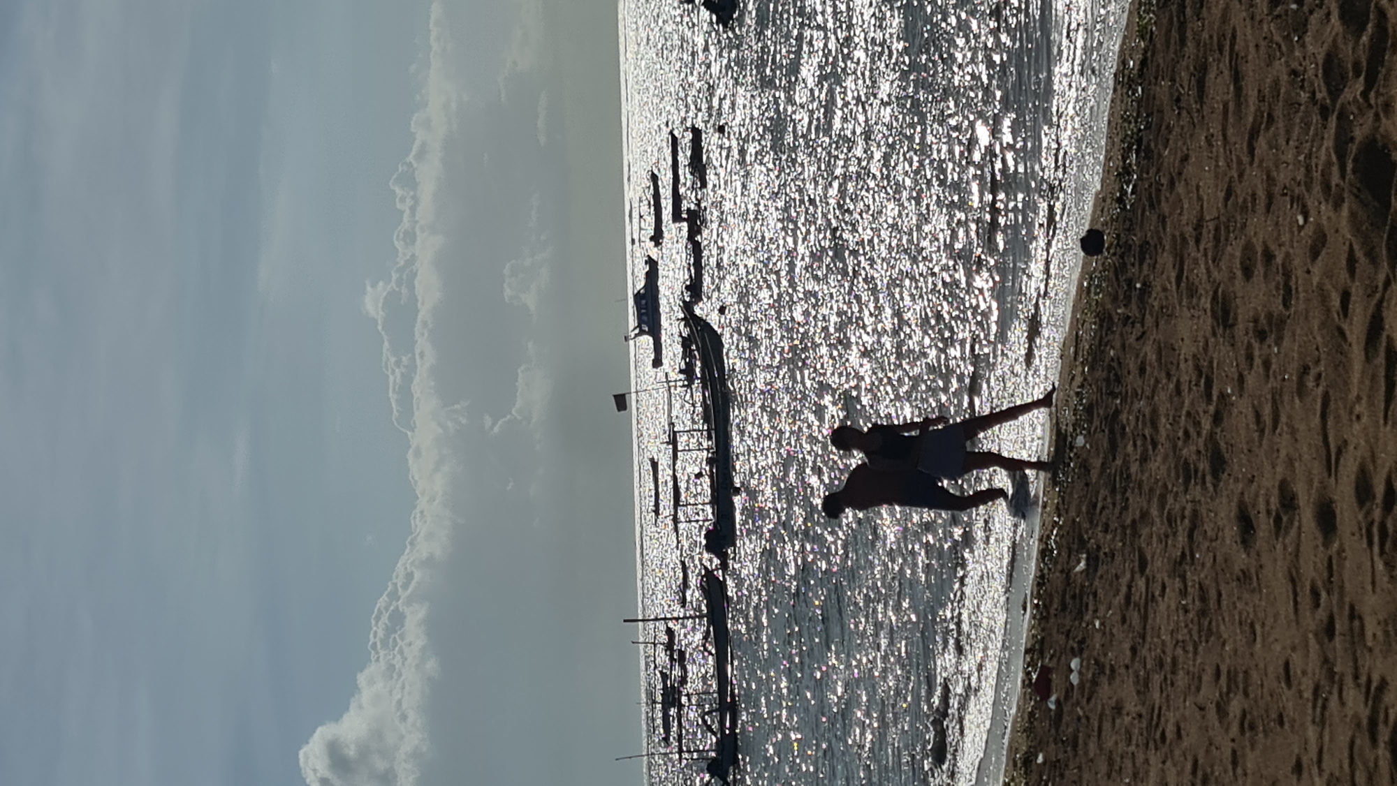 The image depicts a beach scene in Denpasar, Indonesia, with a wide expanse of shimmering ocean in the background. The sun is setting, casting a brilliant glow on the water's surface, creating a dazzling effect. Several boats are anchored offshore, some appearing to be traditional fishing vessels and others motorboats. In the foreground, two individuals are walking along the shoreline, silhouetted against the bright water. The beach itself is sandy, with some debris scattered near the water's edge. The sky above is partly cloudy, with dramatic cloud formations adding to the atmospheric quality of the scene.