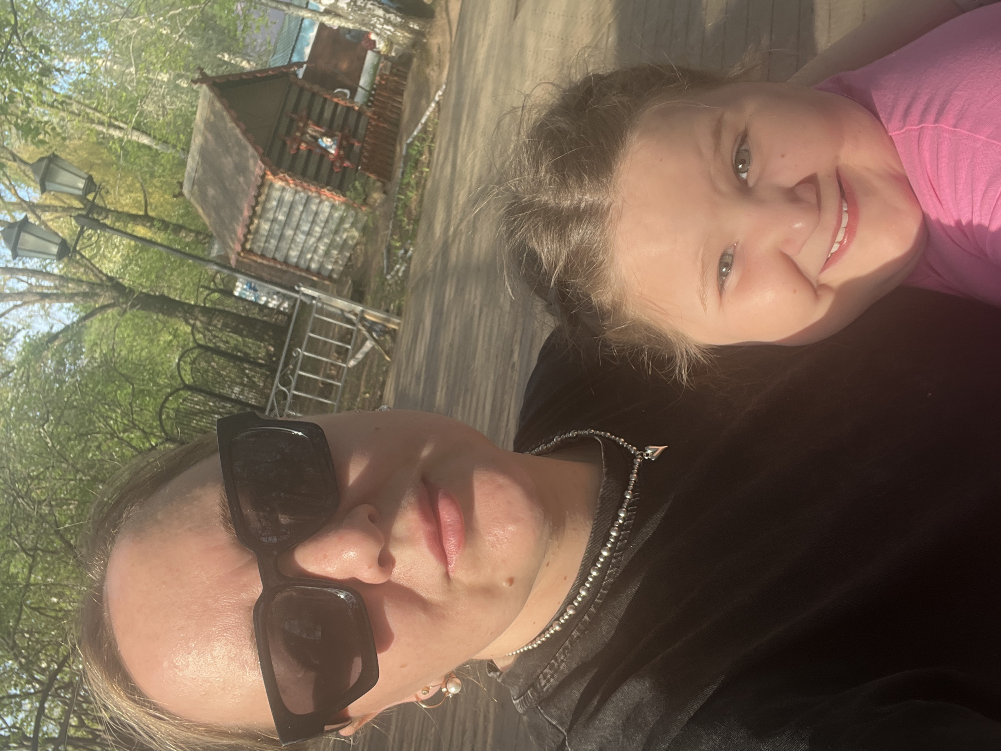 The image shows a woman and a young girl in an outdoor setting in what appears to be a park or recreational area. The woman is wearing sunglasses and a black shirt, while the girl is wearing a pink shirt. The background includes trees, a small wooden structure resembling a house, and a streetlight. This photo was taken in the city of Chaikovsky, Russia. The individuals are likely posing for a selfie or photo.
