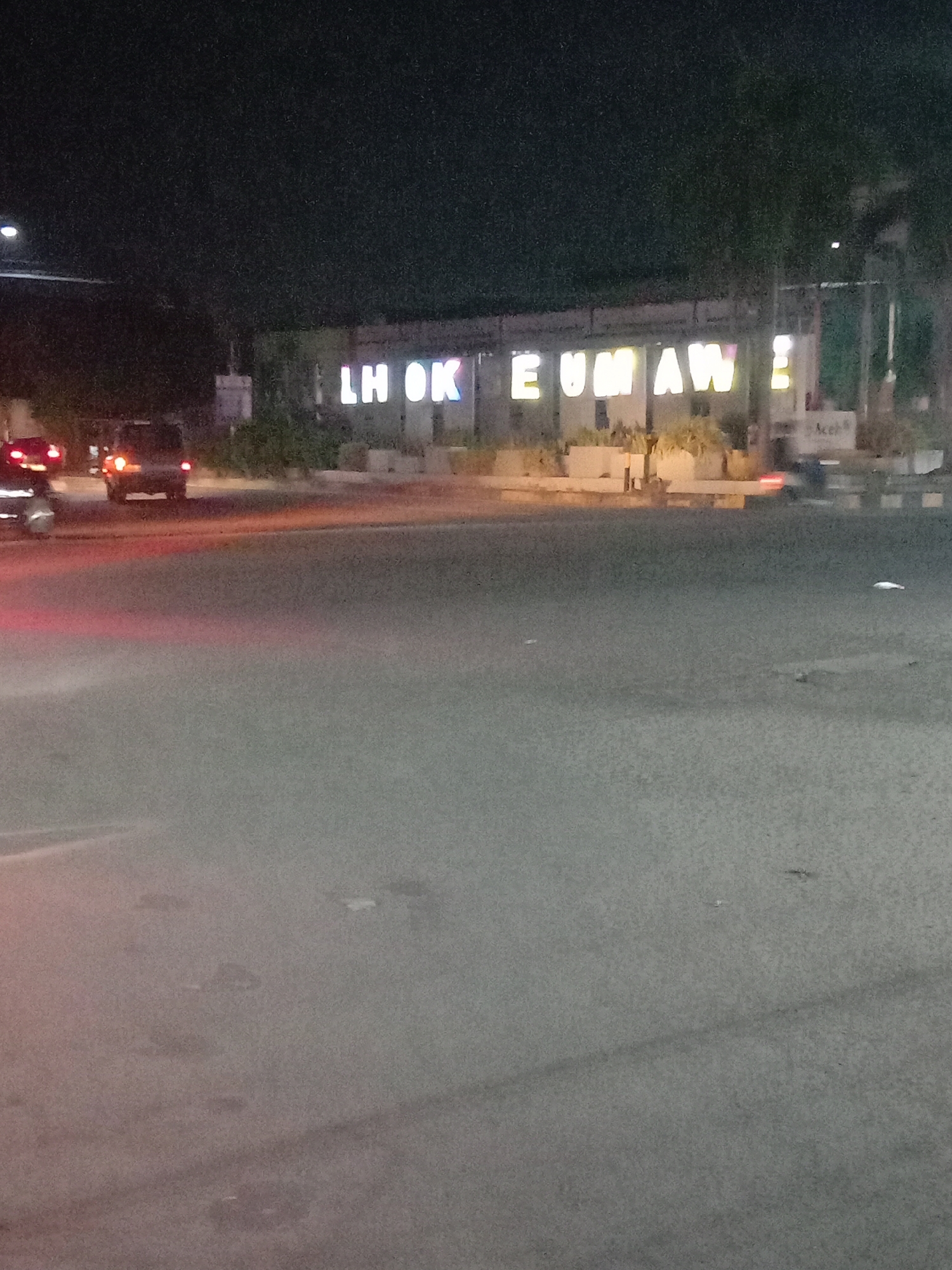 The image captures a nighttime street scene in Lhokseumawe, Indonesia. The foreground is dominated by a dark asphalt road, showing faint light trails from moving vehicles and subtle textures on the surface. In the left midground, several vehicles are visible, primarily indicated by their illuminated red taillights, suggesting traffic or a junction. One dark-colored car with distinct red taillights is clear, moving away from the viewer.
In the background, a prominent, brightly illuminated sign spells out "LHOKSEUMAWE" in large, white capital letters. This sign appears to be affixed to a structure, possibly a building or a decorative wall. Below the sign, there are landscaped areas with planters and some small trees or shrubs. To the right of the "LHOKSEUMAWE" sign, a smaller sign or banner can be seen with the text "Acer". The sky is dark, indicating it is night, and there are no discernible people in the frame. The scene portrays an urban environment with artificial lighting defining the main features.