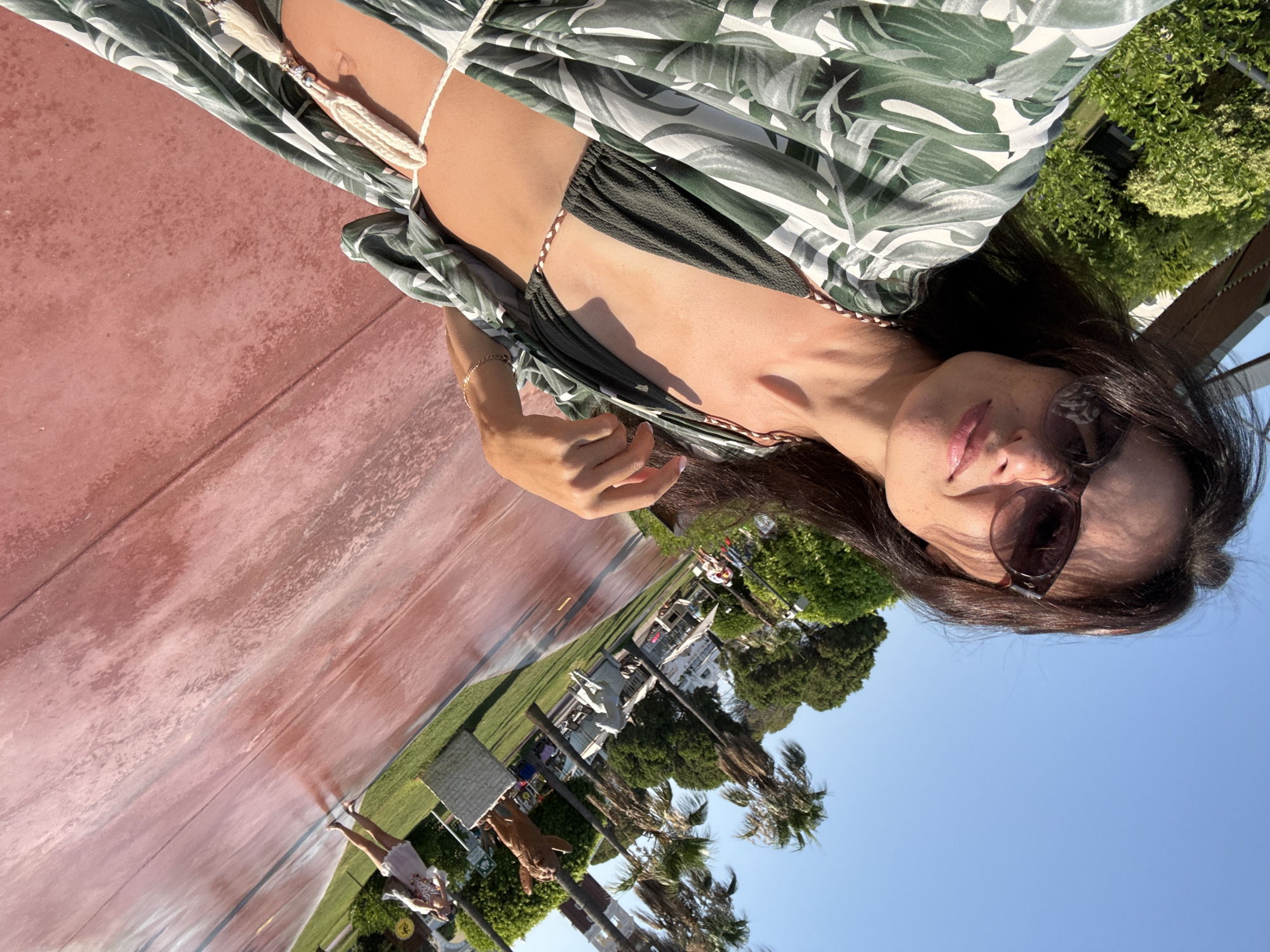 In Aksu, Turkey, on a sunny day, a woman in sunglasses and a green leaf-print cover-up over a dark bikini poses for a selfie. She is standing on a reddish-pink paved walkway that is wet in places, reflecting the bright blue sky and palm trees. Behind her, a lush green garden area is visible, with more palm trees and some structures that appear to be part of a resort or park. In the background, other people can be seen walking and enjoying the outdoor space, including a woman in a light-colored dress. There are also decorative sculptures in the grassy area. The scene suggests a leisurely vacation or holiday atmosphere.