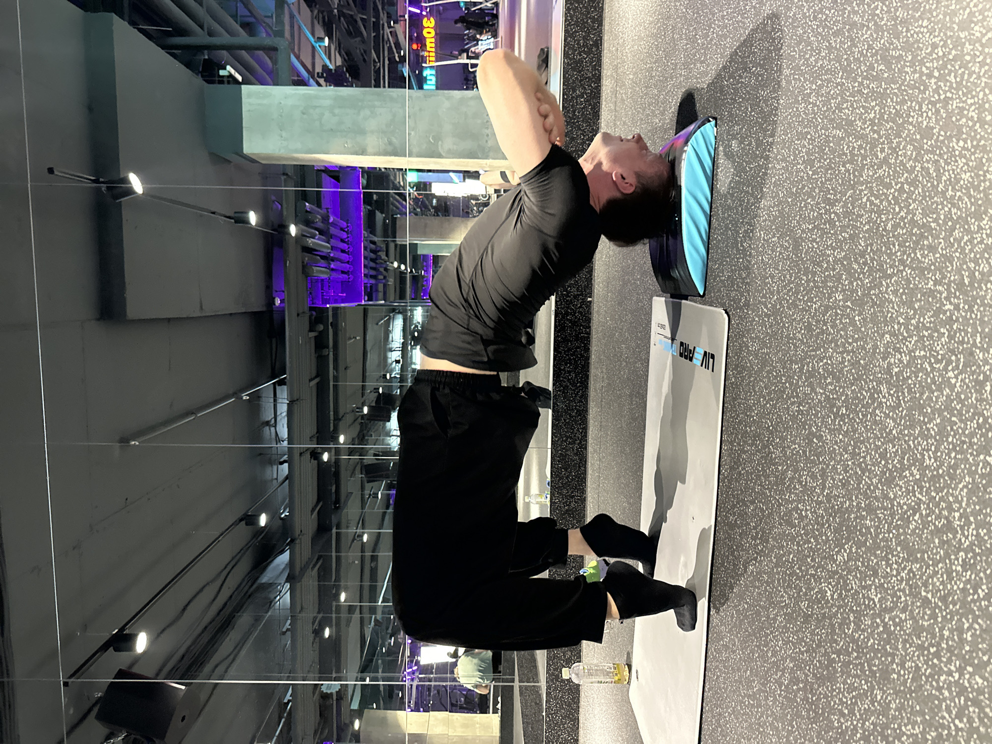 A person is performing a yoga pose in a gym. The individual is wearing black athletic clothing and black socks. They are positioned on a yoga mat, with their head resting on a blue yoga pad. Their body is arched backwards, with their feet on the floor and their hands supporting their head. The background shows a gym environment with mirrors, lights, and purple accents. A water bottle is visible on the yoga mat. The flooring is a speckled grey material.
