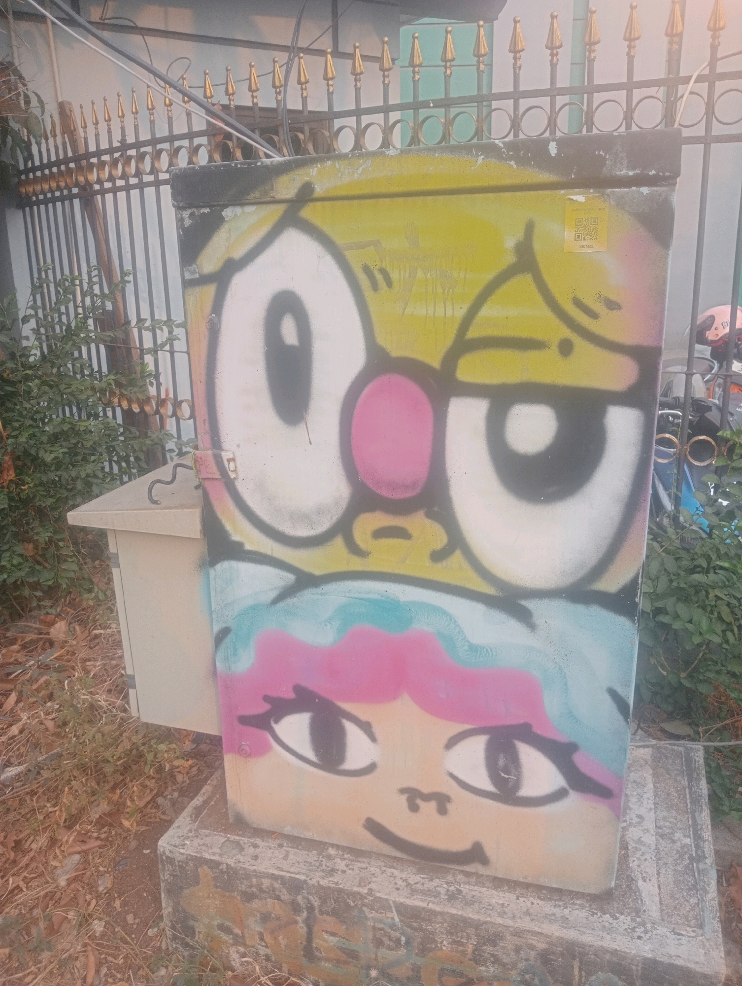 The image shows a street-level view of a graffiti-covered electrical box in East Jakarta, Indonesia. The graffiti depicts two cartoon faces, one with large eyes and a yellow head, and another with blue and pink hair. The electrical box is positioned on a concrete base, surrounded by dry leaves and vegetation. A black metal fence with golden finials runs along the top of the frame, behind the electrical box. In the background, a motorcycle with a pink helmet is visible behind the fence. The scene appears to be outdoors, under overcast skies.