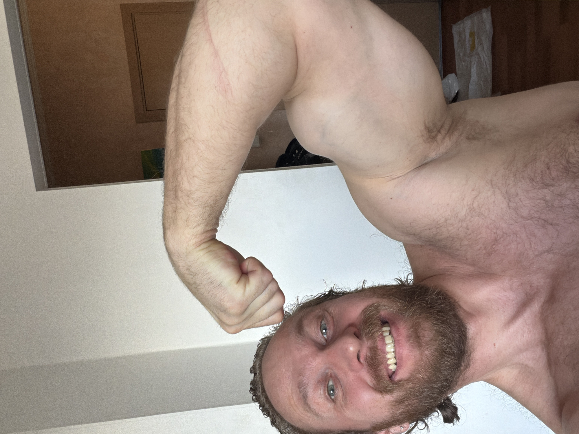 A man is flexing his right bicep and smiling at the camera. He has a beard and short curly hair. He is shirtless. The background includes a white wall, a door, and some items on the floor. The location is not specified, and there is no visible indication it is in Saint Petersburg, Russia.