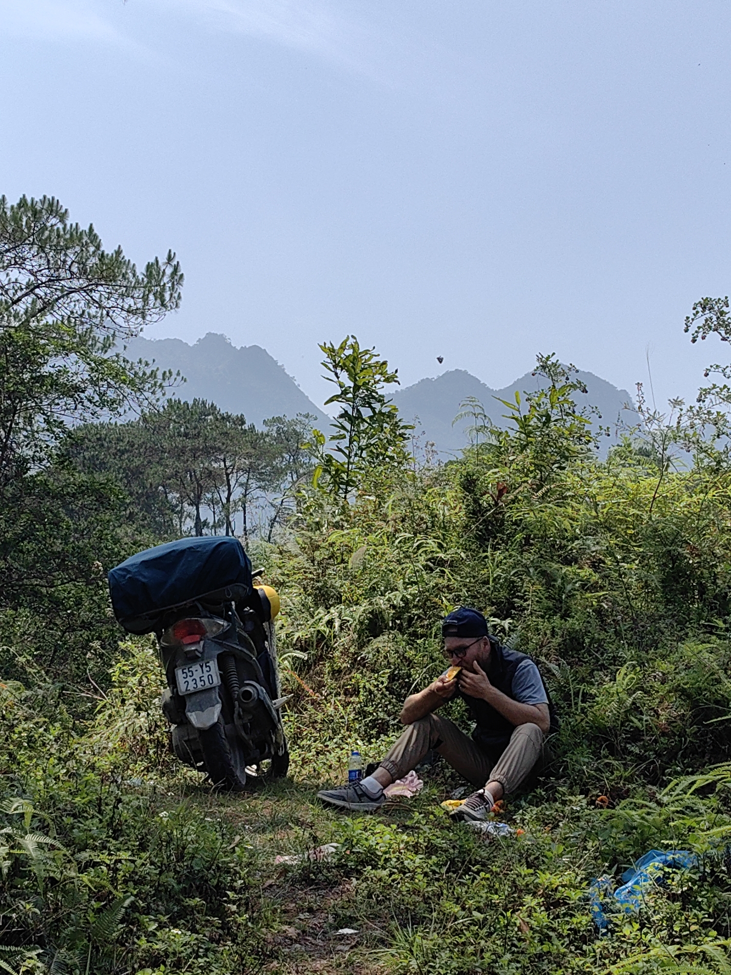 A man is sitting on the ground eating, next to a motorcycle, in a verdant, hilly environment in Yên Minh Commune, Vietnam. The man, wearing a blue cap, glasses, a vest, a blue shirt, and brown pants, is eating what appears to be a piece of fruit with both hands. Next to him is a dark gray motorcycle with a blue tarp covering luggage on the back. The motorcycle's license plate is visible, reading "55-Y5 2350". A water bottle is on the ground near the man. The background shows lush greenery, trees, and distant mountains obscured by a light haze. The foreground is filled with dense undergrowth.