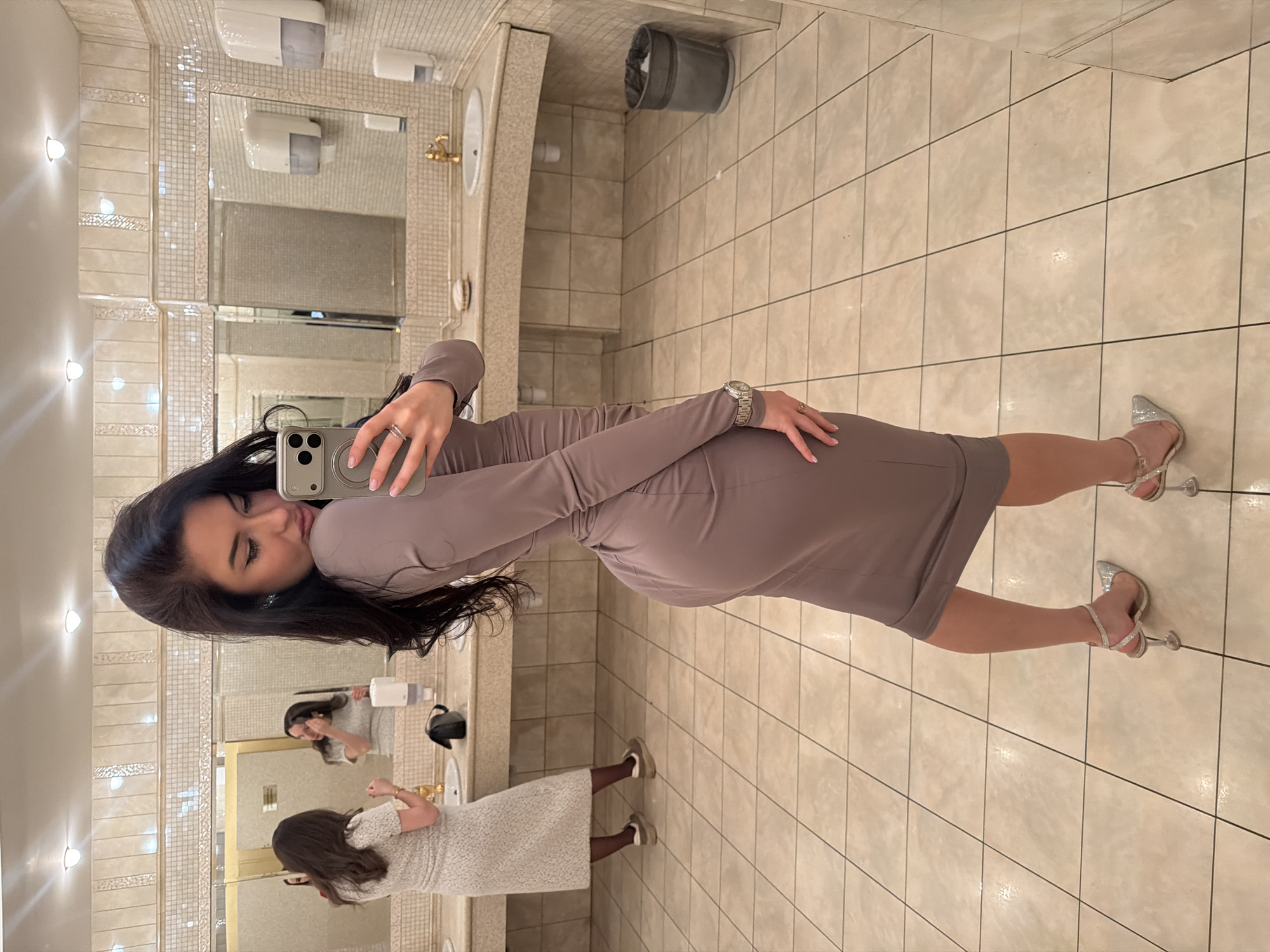 A woman takes a mirror selfie in what appears to be a well-appointed public restroom in Moscow, Russia. The image is oriented sideways, with the woman standing perpendicular to the floor, facing left.
The main subject is a woman with long, dark hair, dressed in a form-fitting, long-sleeved, light brown or taupe dress. She holds a silver-colored smartphone with a prominent camera bump in her right hand, aiming it towards the mirror to capture her reflection. She wears multiple rings on her fingers and a silver-toned watch on her left wrist. Her expression is a slight pout, common for selfies. Her feet are adorned with sparkly silver high heels with ankle straps.
The restroom features light-colored tiled walls, some sections with decorative mosaic-like strips. Bright sconce lights illuminate the area. A large mirror stretches across the wall above a vanity counter with two sinks. Gold-colored faucets are visible on the counter. Various restroom fixtures are mounted on the wall, including white soap dispensers and a hand dryer. A dark gray trash can is positioned next to one of the sinks. The floor is also tiled in a light, neutral color.
In the reflection of the mirror, another woman is visible in the background, further into the restroom. She has dark hair and is wearing a light-colored, possibly lace, dress and dark shoes, appearing to be adjusting something or looking into the mirror as well. The overall setting suggests an upscale public facility, such as a hotel, restaurant, or event venue. There is no discernible text visible in the image.